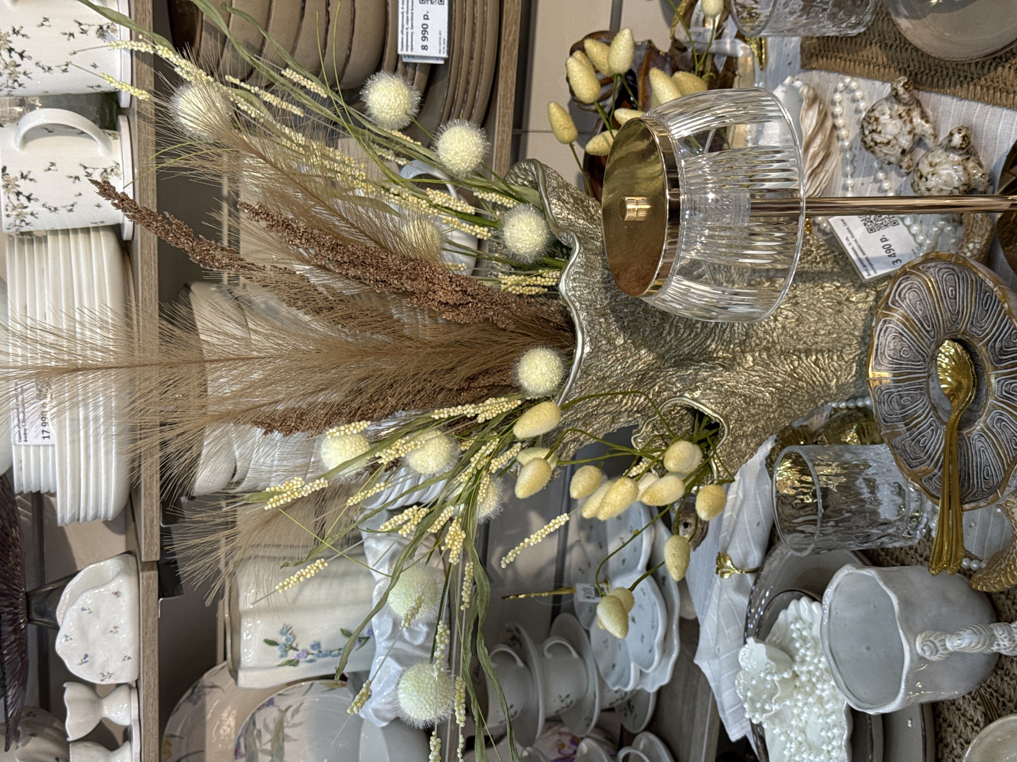 The image depicts a display of tableware and decorative items in a retail setting. The location is likely a store in Krasnoyarsk, Russia, based on a price tag in Russian currency (rubles). The scene features a variety of plates, cups, and mugs, some stacked and others displayed individually. The color scheme is predominantly white, beige, and gold. Dried floral arrangements, including pampas grass and other textured plants, are used as decorative elements, adding height and visual interest to the display. A textured glass object with gold accents is also visible. The overall impression is one of a carefully curated home decor or tableware collection.
