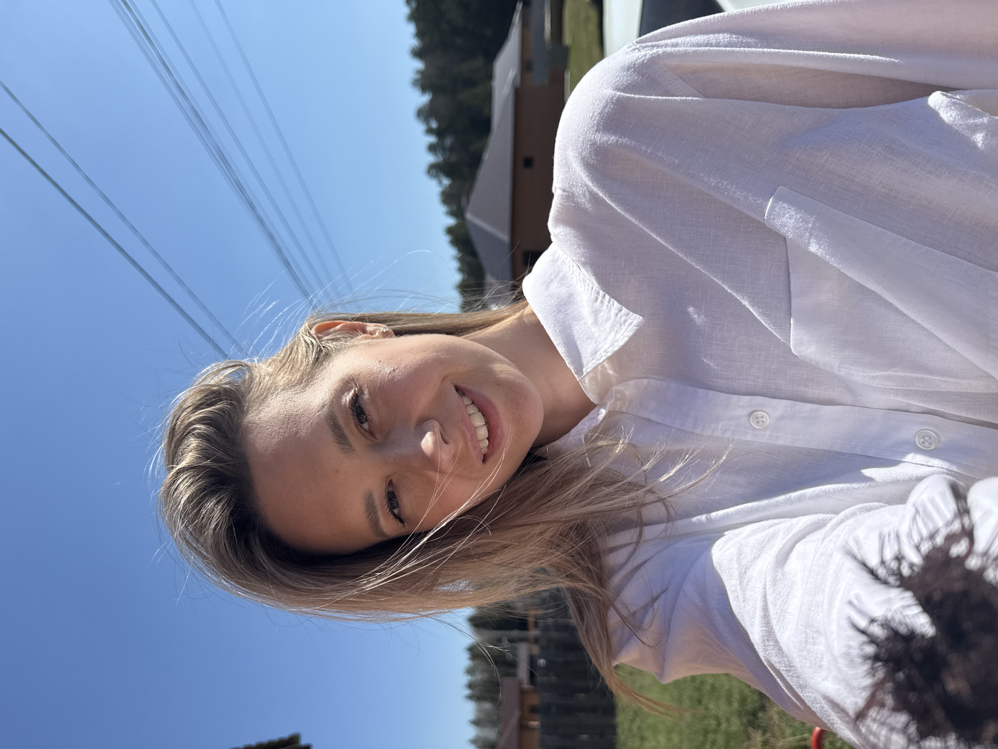 A woman with blonde hair is smiling and looking at the camera. She is wearing a white button-down shirt. In the background, there is a clear blue sky with power lines visible, as well as a building and trees. Based on the location inferred, this scene is likely in a residential area of Chaikovsky, Russia.
