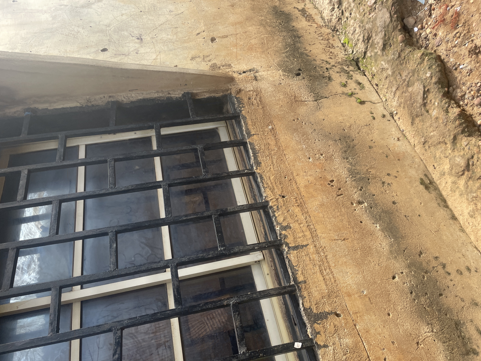 A barred window on the exterior of a building in Lokoja, Nigeria, is captured in a medium-angle, eye-level shot. The window features black metal bars with a grid pattern, offering a glimpse of the dark, reflective glass behind them. The building's wall, made of rough, textured concrete, shows signs of wear, including stains and small holes. The wall is painted a light brown, with darker patches of grime and dirt accumulated near the window and on the lower sections. The ground in front of the wall consists of a rough, uneven concrete surface interspersed with small stones and patches of dirt. Some sparse green moss is visible growing in the cracks of the concrete near the base of the wall. The lighting suggests it is daytime, with shadows indicating the direction of the sun.