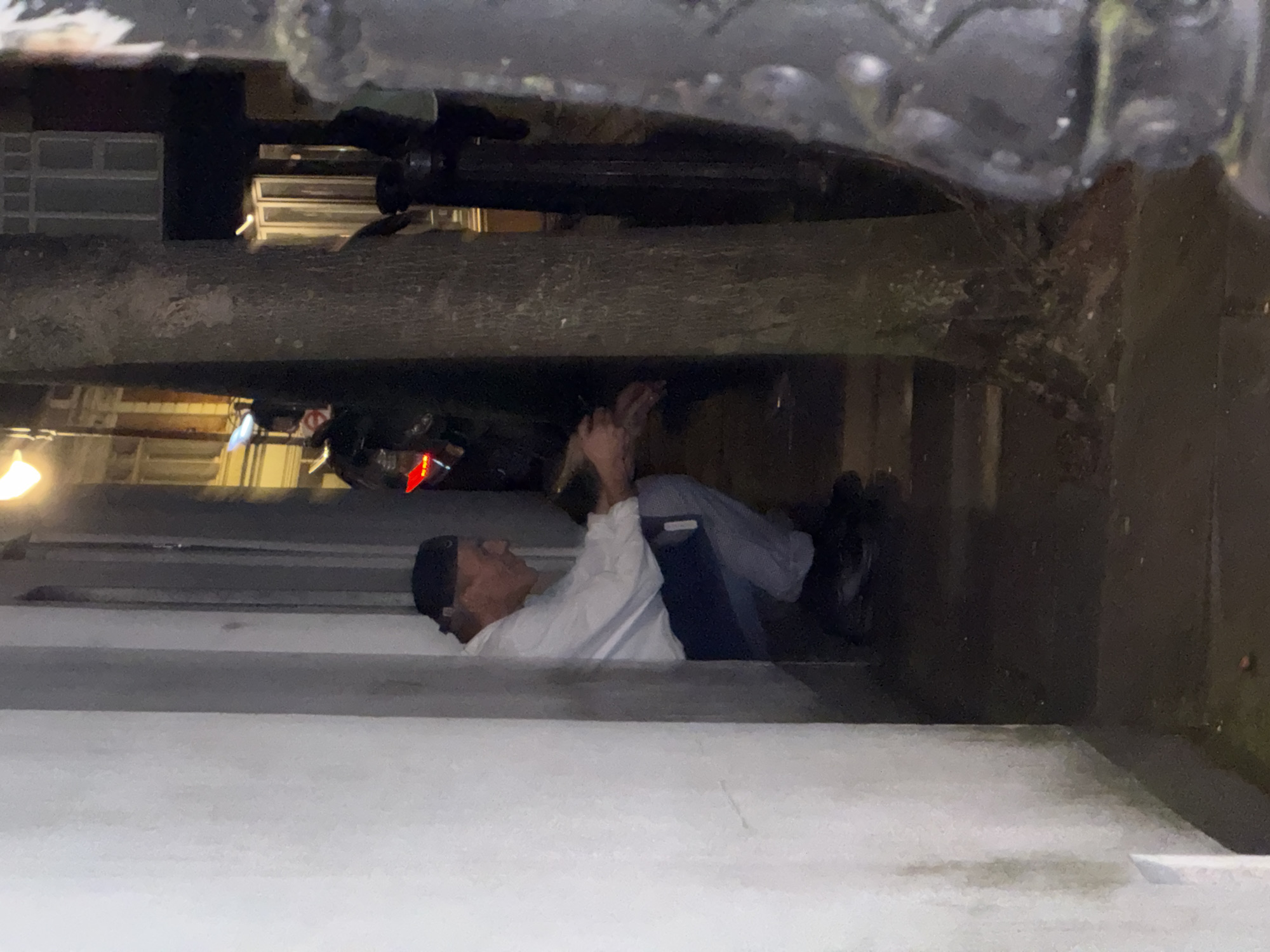This image depicts a nighttime urban scene in London, United Kingdom, within a narrow alleyway or recessed architectural space. In the left foreground, an adult male, appearing middle-aged, is crouched against a light-colored, streaked wall. He wears a dark baseball cap reversed, a white long-sleeved shirt, light-colored trousers (possibly grey or light blue), and dark shoes. His hands are clasped in front of him, and his head is bowed. The ground beneath him is dark and appears damp or wet, reflecting light.
A prominent tree trunk stands centrally to the right, partially obscuring the background. To the far right, a dark, ornate metal street fixture is visible. In the deeper background, illuminated building facades with multi-pane windows are discernible, suggesting a typical London street. Blurred figures are partially visible behind the tree trunk, one wearing a light-colored top. A distinctive red light source is present in the mid-ground, partially obscured. The scene is illuminated by artificial light sources, including bright streetlights at the top left and in the background, casting a yellow-orange glow.