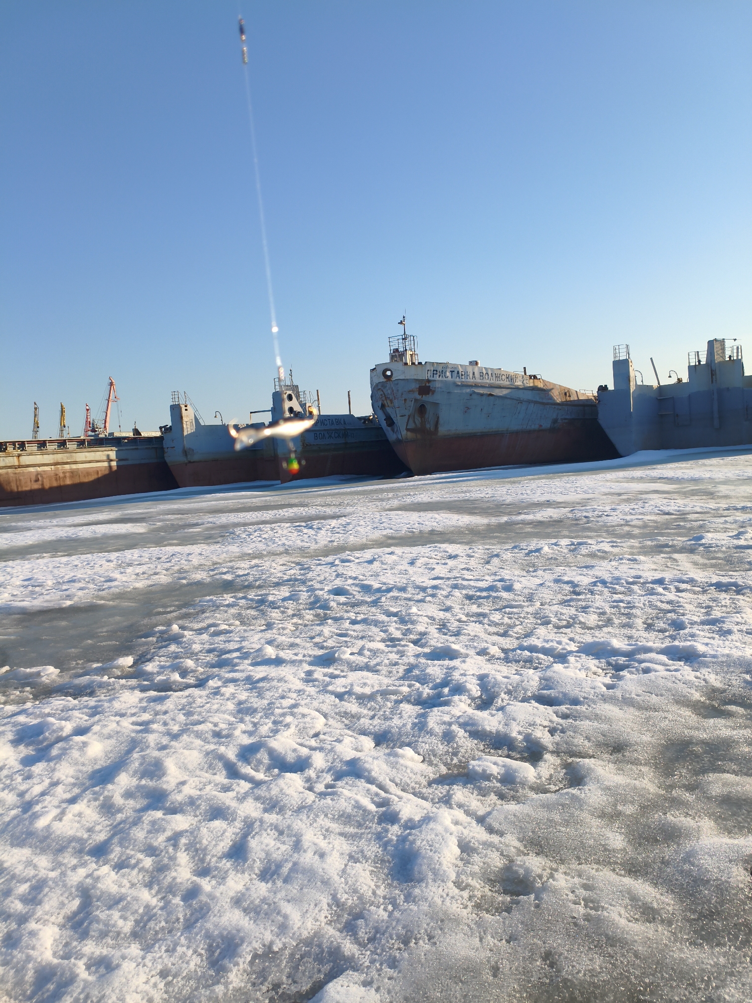 The image depicts several barges frozen in ice at a location in Chaikovsky, Russia. The barges appear to be old and possibly disused. One barge is labeled "ПРИСТАВКА ВОЛЖСКИЙ". The ground is covered in ice and snow. The sky is clear and blue. Some cranes are visible in the background.