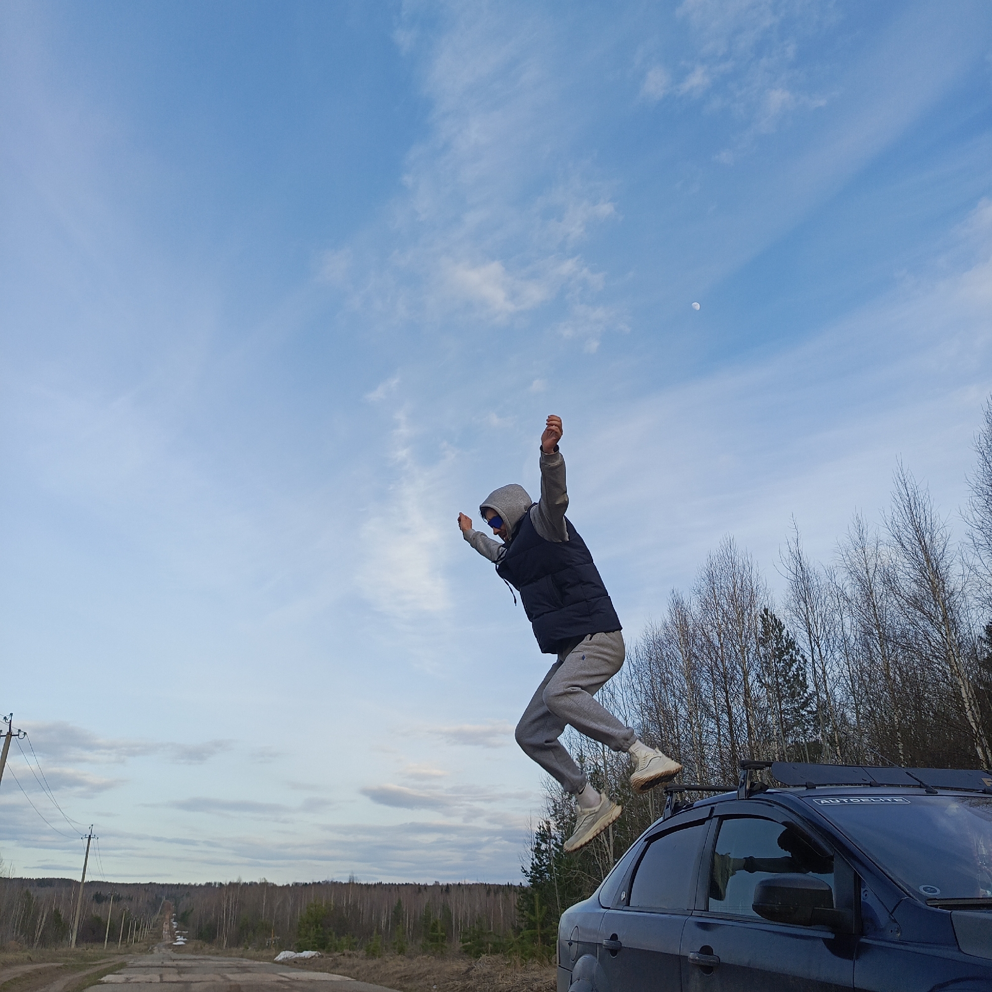 A person is in mid-air, leaping above a dark blue car. The person is wearing a grey hooded sweatshirt, a dark blue vest, grey sweatpants, and light-colored sneakers. The car has roof racks with a sticker that reads "AUTODELITE." The background shows a dirt road leading into a wooded area under a blue sky with scattered clouds and a faint moon. Power lines are visible on the left side of the road. The location is consistent with a rural setting near the city of Markovsky, Russia, as suggested by the scenery and infrastructure present.