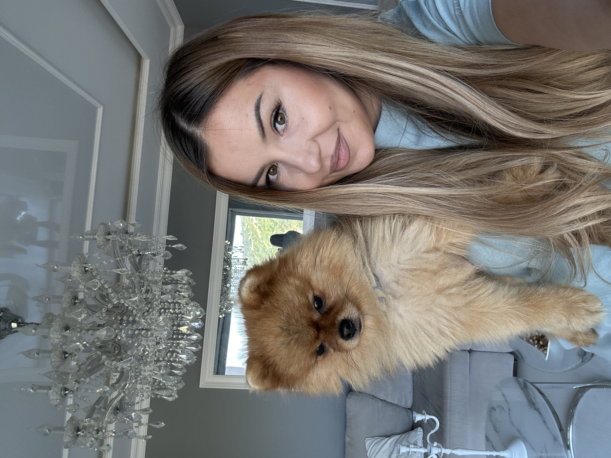 A woman with long, blonde-brown hair and makeup is holding a fluffy, light brown Pomeranian dog. She is wearing a light blue turtleneck shirt. The dog is looking down with its ears slightly back. Behind them, a large crystal chandelier hangs from a gray, coffered ceiling. A window with a view of hills and trees is visible in the background, with a reflection of a chandelier and the woman. Furniture, including a grey sofa and a white candelabra on a marble-top table, is present in the room. The scene appears to be indoors in a well-decorated living space. The location is Asenovgrad, Bulgaria.