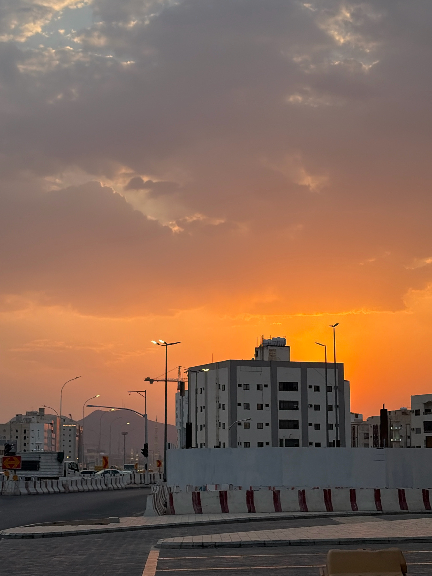 This is a photograph of a street scene in Medina, Saudi Arabia, at sunset. The sky is a vibrant gradient of orange and pink, with dramatic clouds illuminated by the setting sun. In the foreground, a street is visible, bordered by concrete barriers with red and white stripes. Several buildings are present, including a modern multi-story white building with dark windows, and other structures further in the distance. Streetlights are visible throughout the scene. A large mountain is visible in the background. The image appears to have been taken from a low angle, looking towards the horizon. There are no people clearly visible in the image.