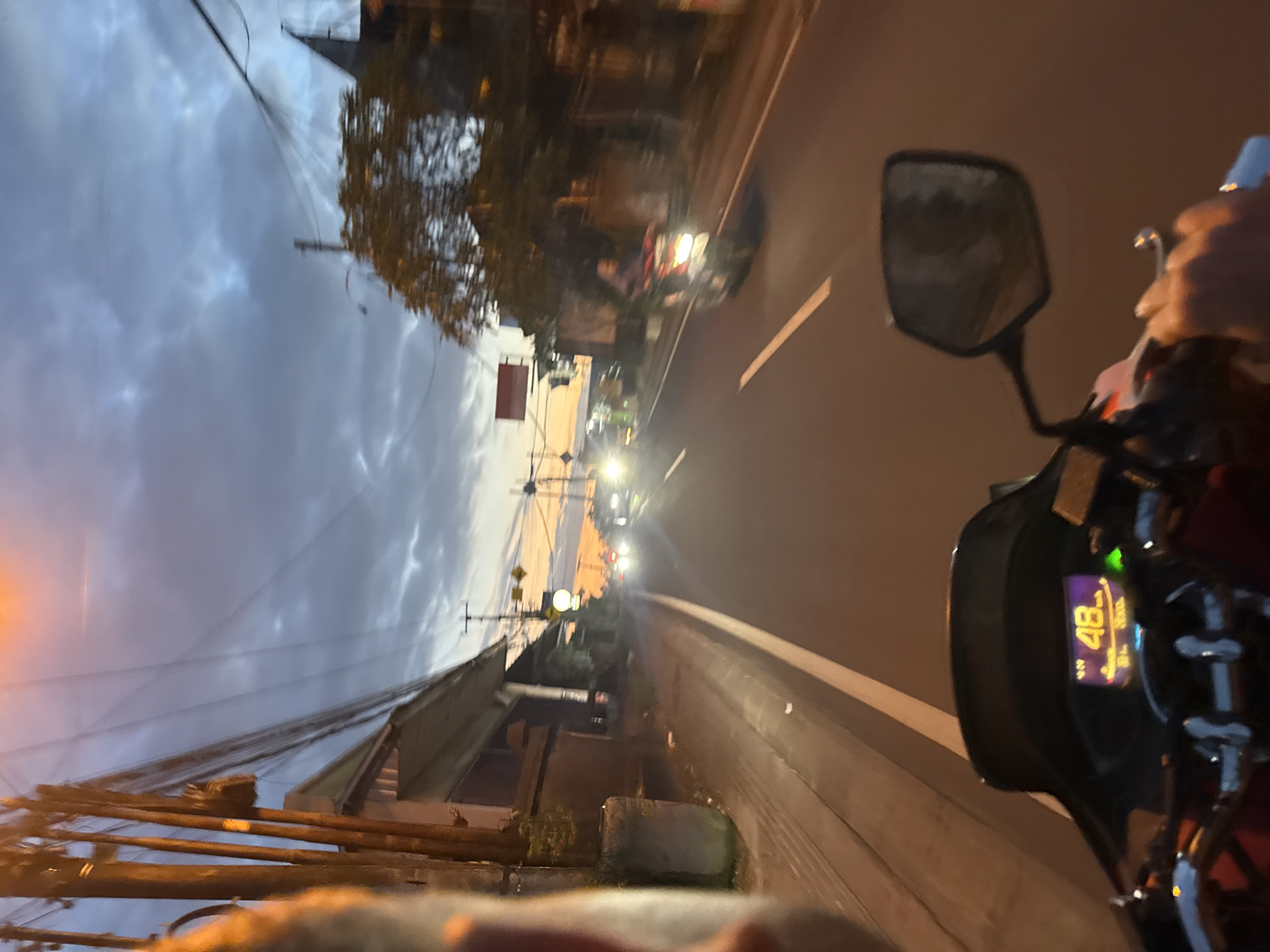 The image depicts a motorcycle riding on a road at dusk. The motorcycle's speedometer reads "48." Another vehicle is visible ahead. The sky is cloudy. The location appears to be a city street, based on streetlights and buildings. It is not possible to confirm this is Kuta Selatan, Indonesia from the image.