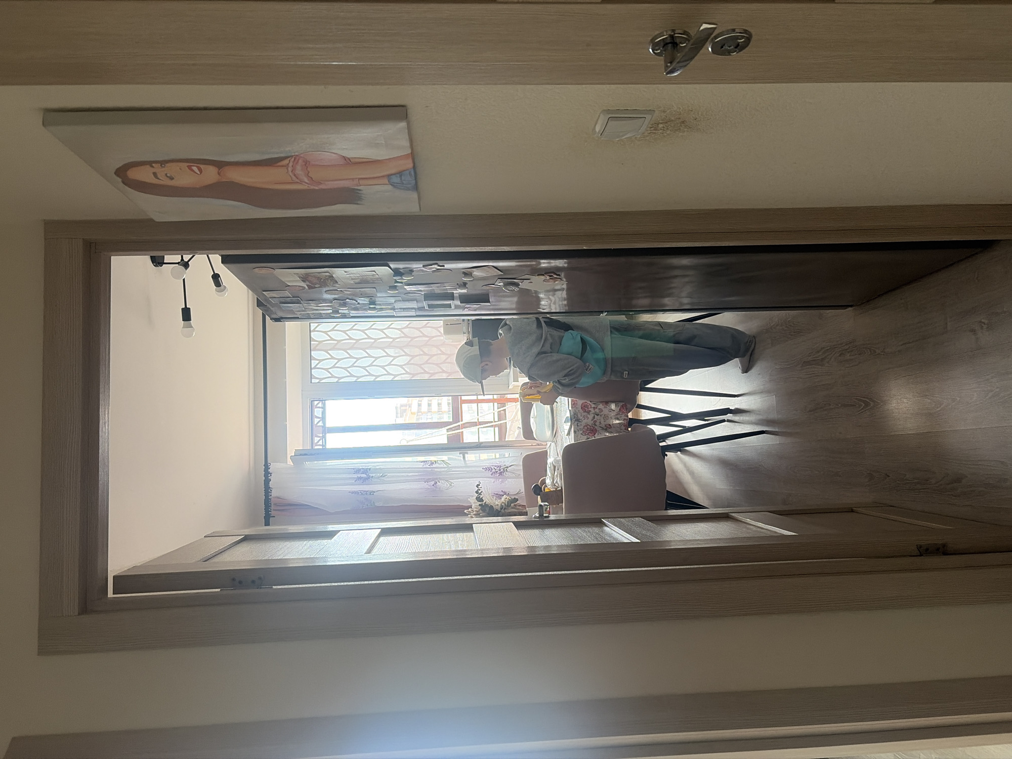 A doorway frames a room. A young boy is visible in the room, wearing a grey hooded sweatshirt and light-colored pants. He stands at a table, and several chairs are nearby. A window reveals a view of a city with buildings, potentially within Saint Petersburg, Russia. Above the doorway, a painting of a woman adorns the wall. A light fixture is partially visible within the room. The floor has a wood-like finish.