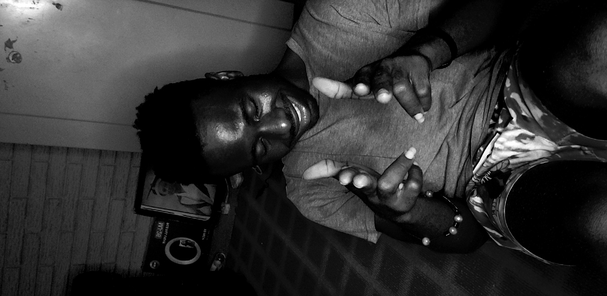 A dark-skinned individual, likely male, is captured in a vertically oriented black and white photograph, appearing to lie down with his head to the left of the frame and body extending to the right. He has short dark hair and is wearing a plain t-shirt and patterned shorts. A black band is visible on his left wrist, and a beaded bracelet with light-colored beads adorns his right wrist.
The person is smiling broadly with eyes closed, conveying a sense of joy or relaxation. Both hands are raised towards the viewer, with fingers spread and slightly bent in a playful or expressive gesture.
The scene is set indoors, possibly in a casual home environment like a bedroom or living room, as the individual appears to be lying on a bed or couch. The background features a textured wall or surface, and to the person's left, a box or frame is visible. The lighting is dim, creating prominent highlights on the person's forehead, cheekbones, nose, lips, and hands. The photograph's orientation suggests either the subject was lying sideways relative to a standard upright camera or the camera itself was tilted. The location context is Narayi, Nigeria.
On the box or frame in the background, inverted text is visible, including "WR-SE", "10 INCH", and other text that appears mirrored or stylized, such as "BCS HIFF THCIM".