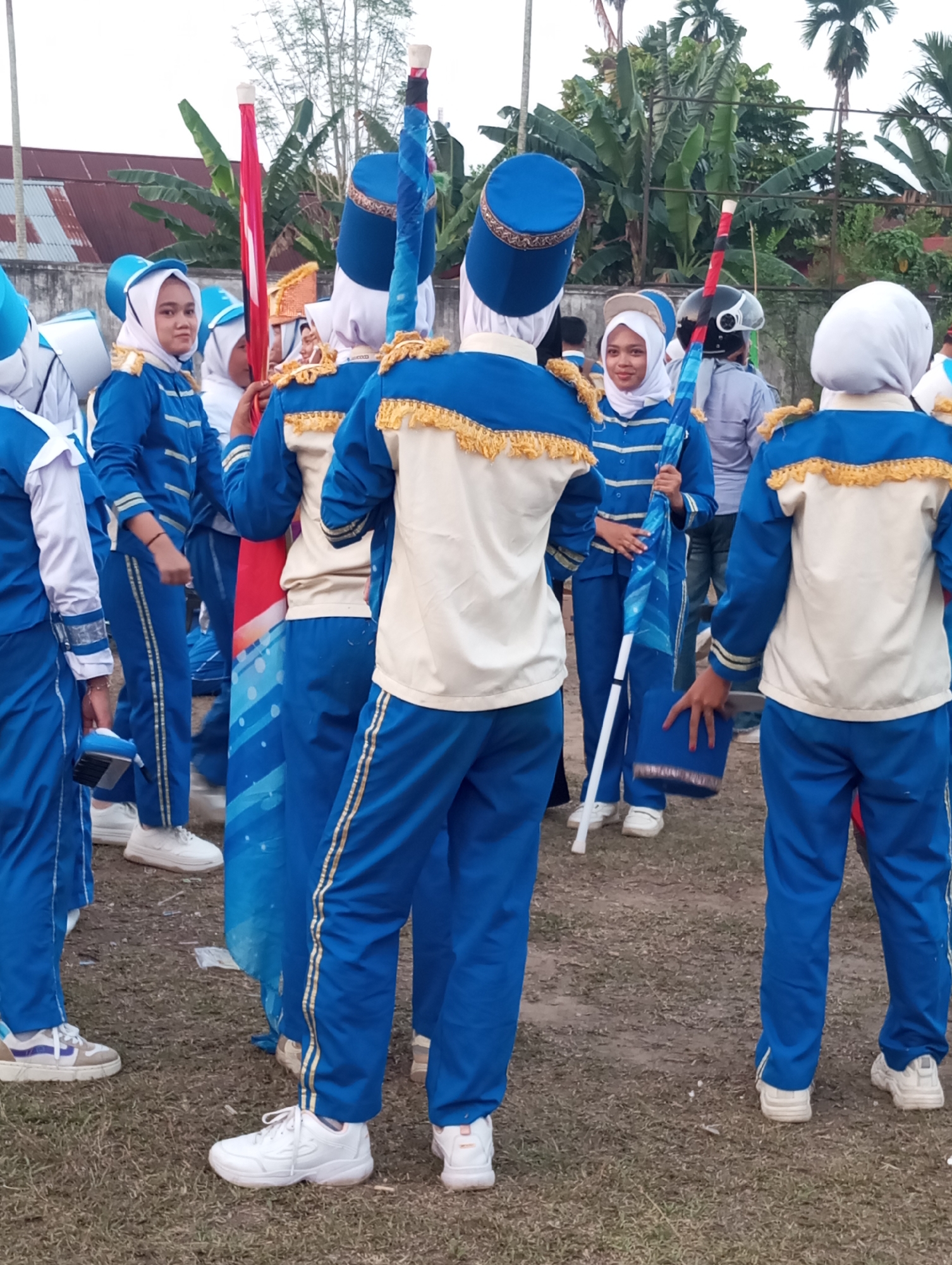 A group of young people in matching blue and gold marching band uniforms are gathered outdoors on a dirt and grass surface. The uniforms consist of blue jackets with gold trim on the shoulders and cuffs, and matching blue pants with gold stripes down the sides. Several individuals are wearing white headscarves. Some are holding long, cylindrical objects, likely flagpoles or batons, with colorful fabric attached. One person is holding a blue cylindrical object with a gold trim, possibly a drum or part of a uniform accessory. In the background, there are green trees, a building with a corrugated metal roof, and a cloudy sky. The scene appears to be in Candirejo, Indonesia, as indicated by the context provided.