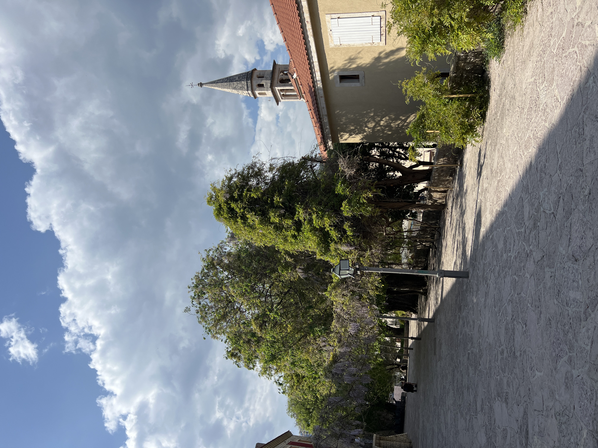The image, rotated 90 degrees clockwise, captures an outdoor scene featuring historic architecture, lush vegetation, and a paved pathway under a dynamic sky.
On the upper right side, a building with a traditional reddish-orange tiled roof and a light-colored, possibly pale yellow, facade is prominent. It features a window fitted with closed white louvered shutters and a smaller, open rectangular window below it. Behind this building, a striking church or bell tower rises, crowned with a pointed spire and topped by a cross or weather vane. The tower exhibits arched openings in its upper section.
The middle and lower sections of the image are dominated by abundant green foliage from several large trees. Hints of purple blossoms, possibly wisteria, are visible among the leaves, and vines cling to some of the trees. A dark street lamp with a square lantern stands amidst the greenery.
The ground, appearing on the left side of the rotated image, is a textured pathway paved with irregularly shaped, light-grey stones. Long, distinct shadows are cast across this paved surface from the direction of the trees and buildings. A small figure in dark clothing can be faintly seen in the distance along the path, suggesting a person walking or resting.
The background, also on the left, showcases a bright sky with patches of blue interspersed with large, fluffy white and grey clouds. The strong sunlight and clear shadows indicate it is daytime, likely morning or late afternoon. The scene evokes a peaceful atmosphere characteristic of an old European town, consistent with the provided context of Budva, Montenegro. No visible text is present.