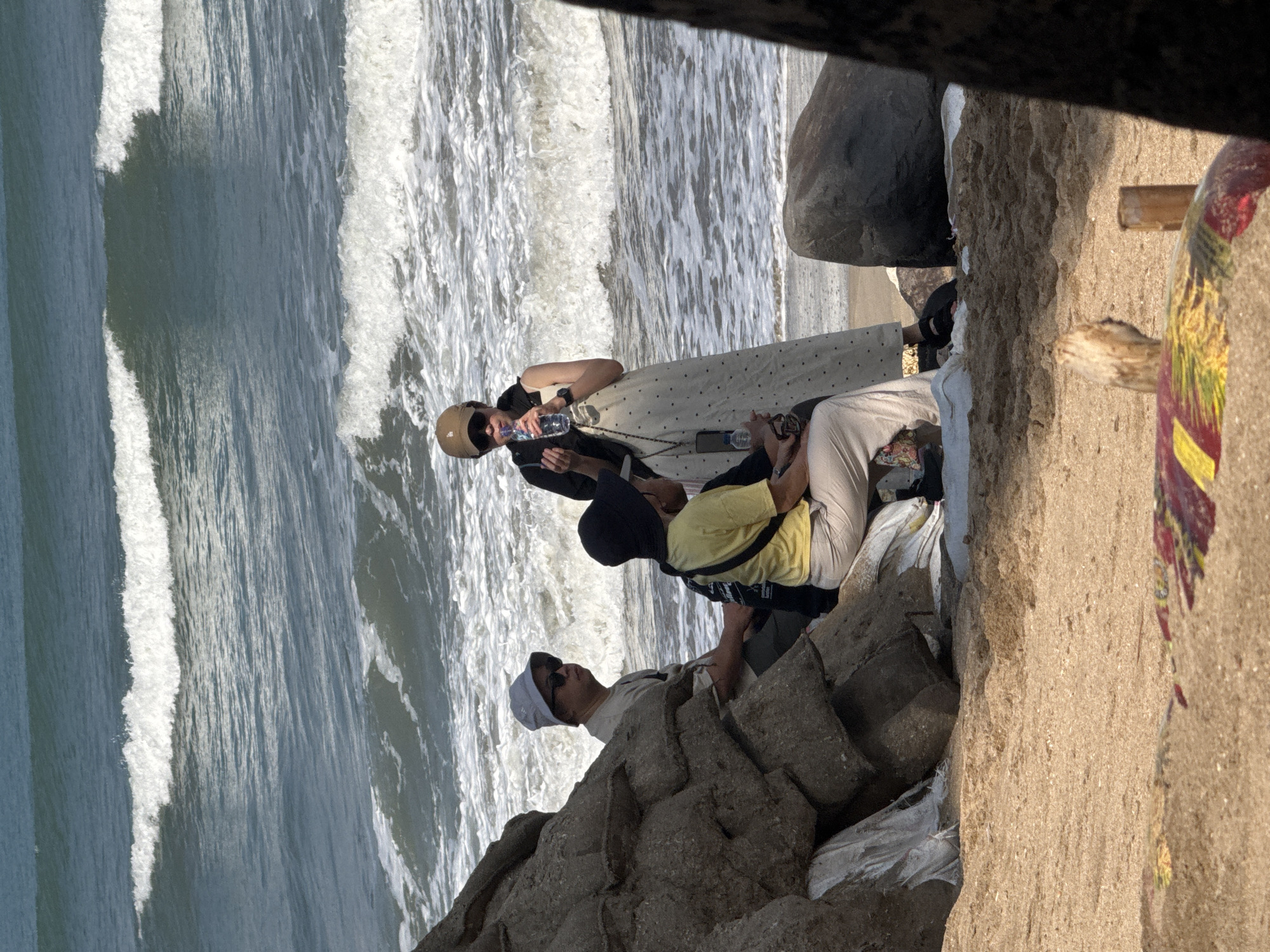 In Kuta, Bali, Indonesia, a group of people are gathered on a sandy beach near large rocks that have been reinforced with white sandbags. The ocean is to the left, with waves crashing onto the shore. In the foreground, a person in a yellow t-shirt and light-colored pants sits on a sandbag, facing away from the camera. Beside them, a person wearing a light-colored t-shirt and a light-colored bucket hat, sunglasses, and a grey t-shirt is seated, looking to the right. Standing to the right of the seated individuals is a person in a cream-colored polka-dot dress, a black jacket, and a brown baseball cap, holding a clear plastic bottle of water and a small object in their hands. A dark shadow on the far right of the image suggests a tree or structure is obstructing the view. The beach is comprised of light brown sand, with some debris visible in the foreground, including a colorful object with red and yellow markings.