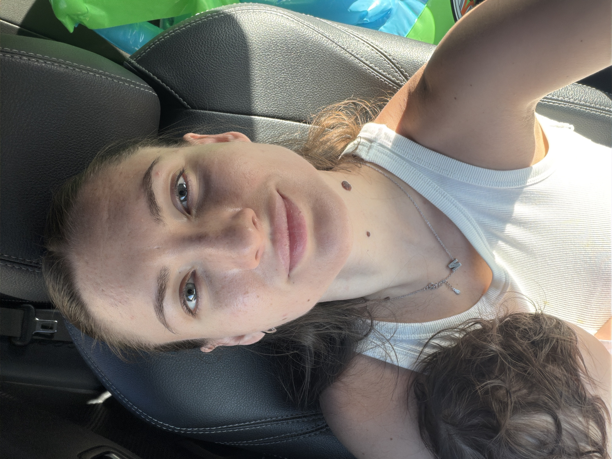 The image shows a close-up, selfie-style shot of a woman sitting in a car seat, likely in Sharm El Sheikh, Egypt, given the context provided. She is wearing a white ribbed tank top and a silver necklace with a pendant. A portion of a child's dark hair is visible on the left side of the frame, suggesting the woman is with a child. The background includes the dark leather interior of the car, with sunlight casting shadows on her face and shoulder. Brightly colored objects, possibly bags or inflatables, are partially visible in the upper right corner. The woman's face is centrally located, with her gaze directed towards the camera.