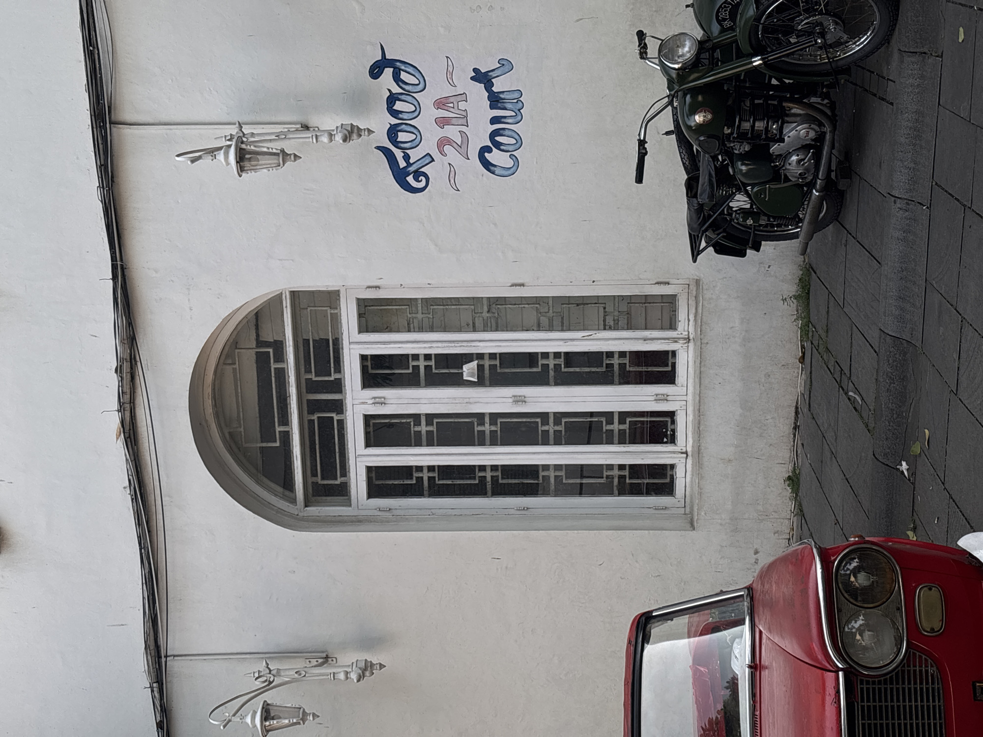 The image depicts the exterior of a building in West Jakarta, Indonesia. The building has a white facade, adorned with a sign that reads "Food 21A Court" in a stylized blue font. Below the sign is an arched window with a white frame and multiple rectangular panes. Attached to the wall are two ornate, white lampposts. Black electrical cables run along the building's left side.
Parked on the sidewalk adjacent to the building is a vintage green motorcycle. The front portion of a red vintage car is also visible, with its distinctive double headlights and chrome grille. The sidewalk is paved with grey stone blocks.