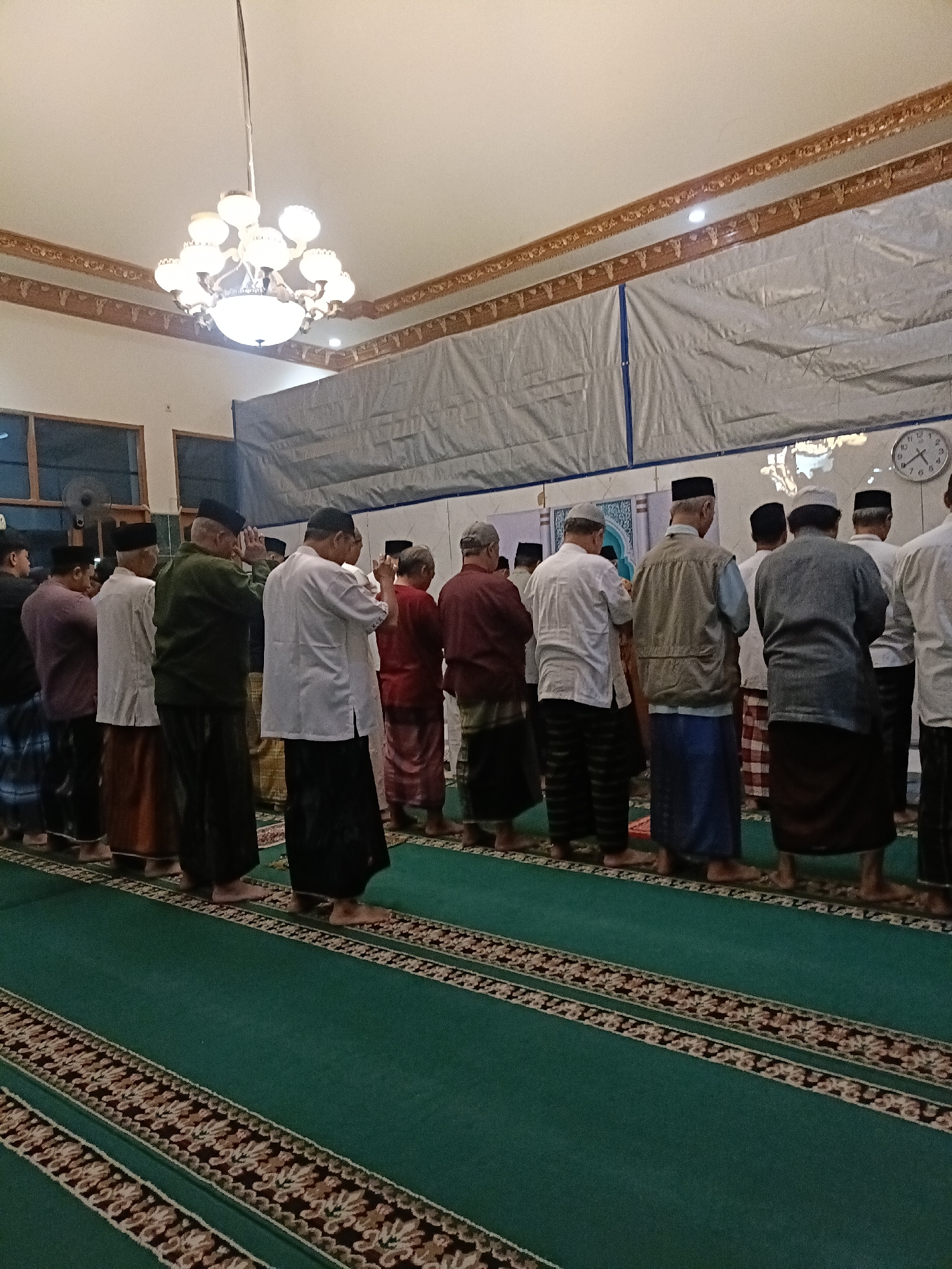 The image shows a group of men standing in a mosque, facing the same direction, presumably in prayer. They are all wearing traditional Islamic attire, including long shirts and sarongs, and some are wearing skullcaps. The mosque has a green carpet with ornate borders, and a chandelier hangs from the ceiling. In the background, a clock is visible on the wall. The men appear to be of various ages and are standing in neat rows. The lighting in the mosque is dim, with the chandelier providing most of the illumination. The overall atmosphere is one of solemnity and devotion. The image was taken in the city of Kediri, Indonesia.