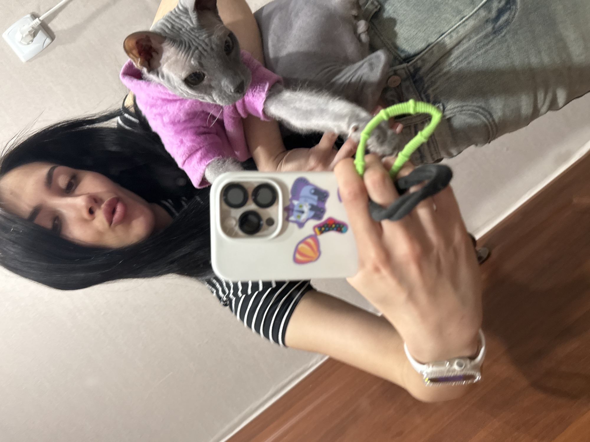 A woman takes a selfie with her hairless cat. The woman has long, black hair and is wearing a black and white striped shirt. The cat is wearing a pink sweater and is being held by the woman. The woman is holding a cell phone with a white case that has stickers on it. The phone has a triple camera array. In the background, there is a white wall and a brown floor. The location is unknown and cannot be determined from the image.