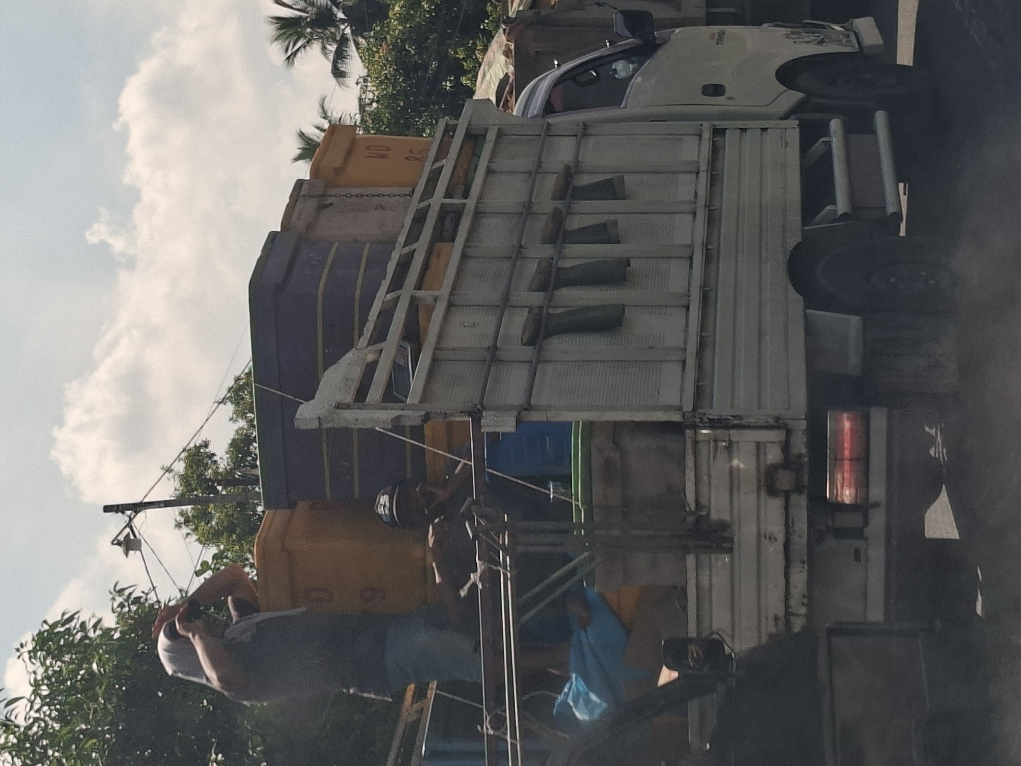 A blurry, medium shot captures a light-colored truck loaded with large yellow and purple containers driving on a road in Lhoknga, Indonesia. The truck has a white cab with a small dark shadow near the driver's side, suggesting a person is inside. In the bed of the truck, a person wearing a dark shirt and a head covering is visible. Another person wearing a dark hat is seated further back, possibly looking at their phone. Several large, rectangular containers are stacked on the truck's flatbed. The surrounding environment shows lush green trees and a cloudy sky with palm trees visible in the distance. The truck's brake lights are illuminated.