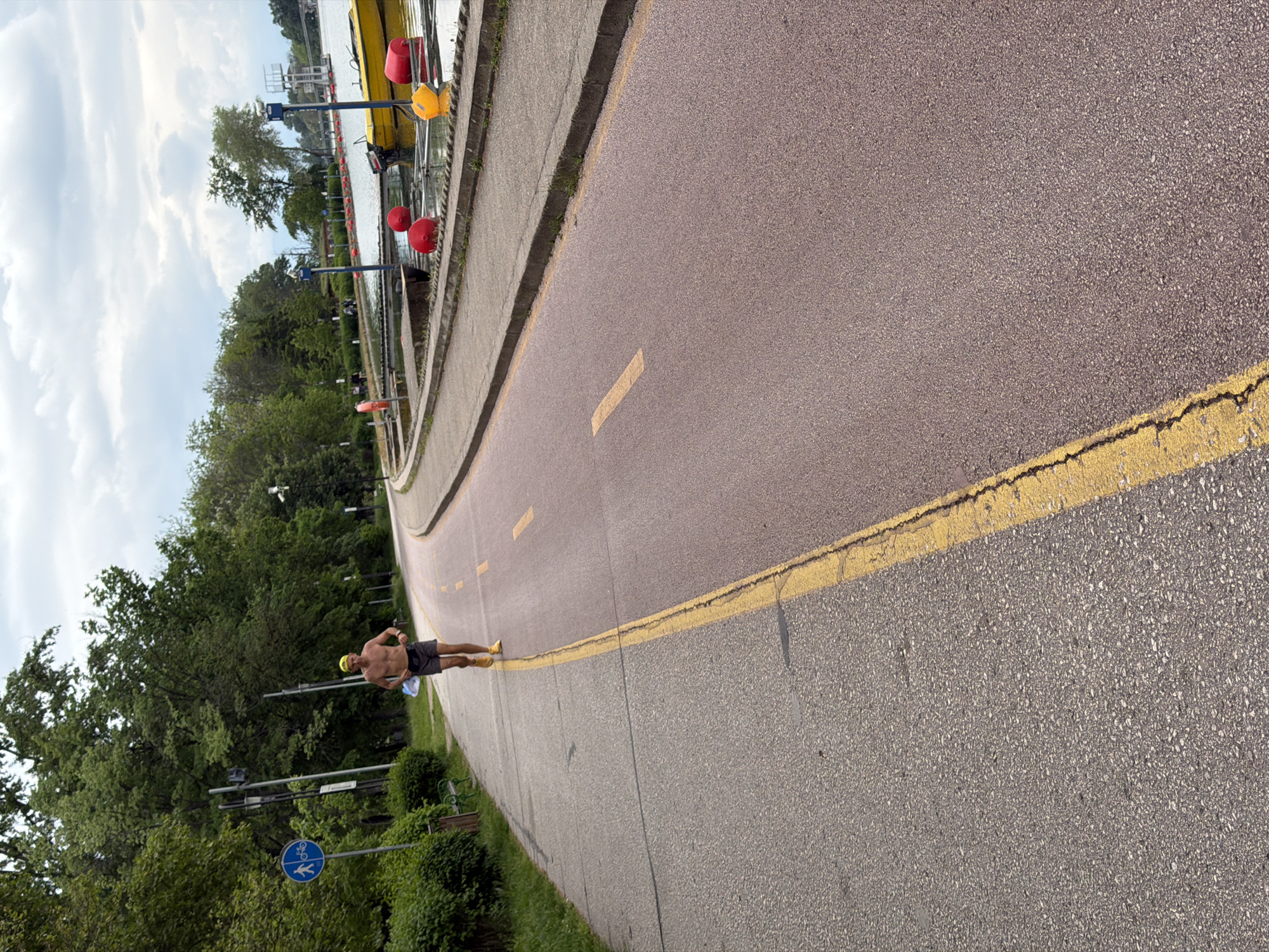 A photograph captures a man jogging along a path in Plovdiv, Bulgaria. He is shirtless, wearing dark shorts, a yellow cap, and yellow running shoes. He carries a clear plastic bottle in one hand. The path is marked with a yellow line and short, dashed yellow lines. To the left, there is greenery and signage, including a blue sign with a white bicycle and pedestrian symbol. To the right is water and a yellow boat. The sky is overcast with white clouds.