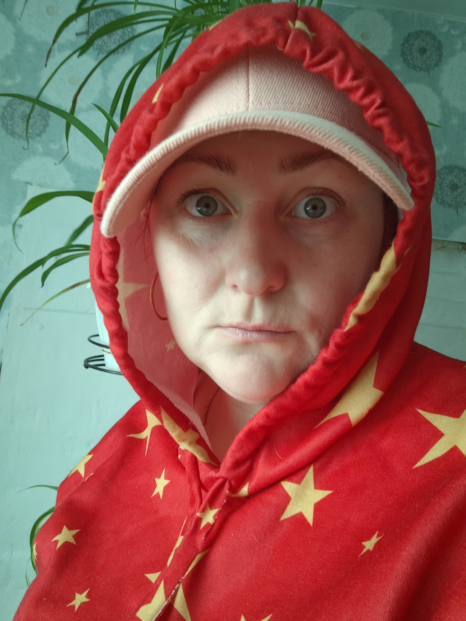 A close-up shot depicts a person with fair skin and gray eyes wearing a pink baseball cap beneath a red hooded sweatshirt adorned with yellow stars. The hood is drawn closely around their face, framing it. They are wearing a hoop earring. The background includes a light blue wall with a plant. The image was taken indoors. The location in the city of Фоки, Russia is not discernible in the image.