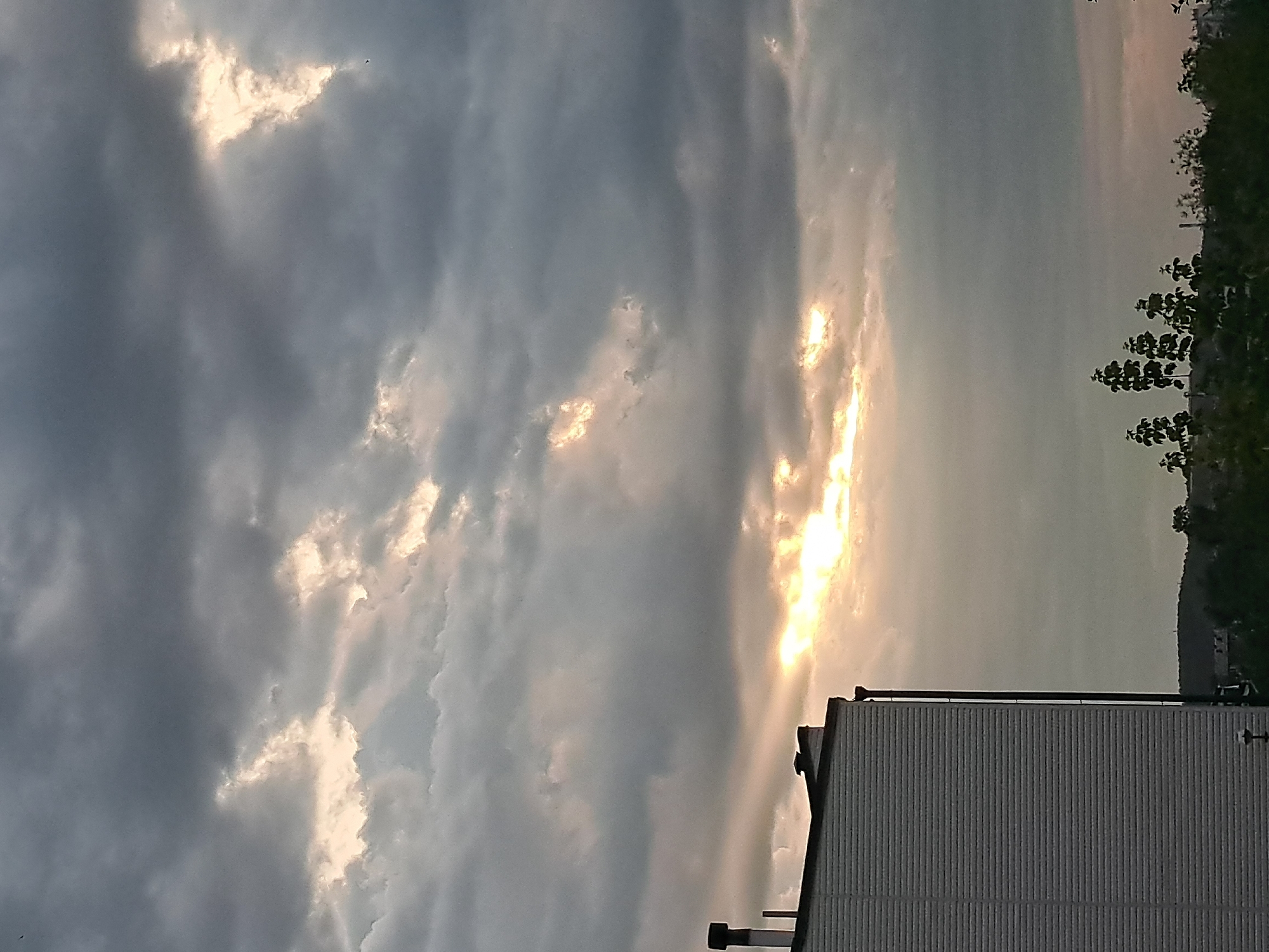 The image shows a cloudy sky with shafts of sunlight breaking through, creating bright patches and illuminating the clouds. In the lower left of the frame, the side of a white building with horizontal siding is visible. A dark chimney pipe extends vertically from the building. In the distance, a treeline with scattered trees is silhouetted against the sky. There are no people visible in the image. The overall mood is atmospheric, possibly indicating an approaching storm or a break in the weather. The scene is set against a backdrop of a possibly urban or suburban environment given the building and distant structures. The lighting suggests it is either dawn or dusk. There is no specific information in the image that would pinpoint a location within Irkutsk, Russia.