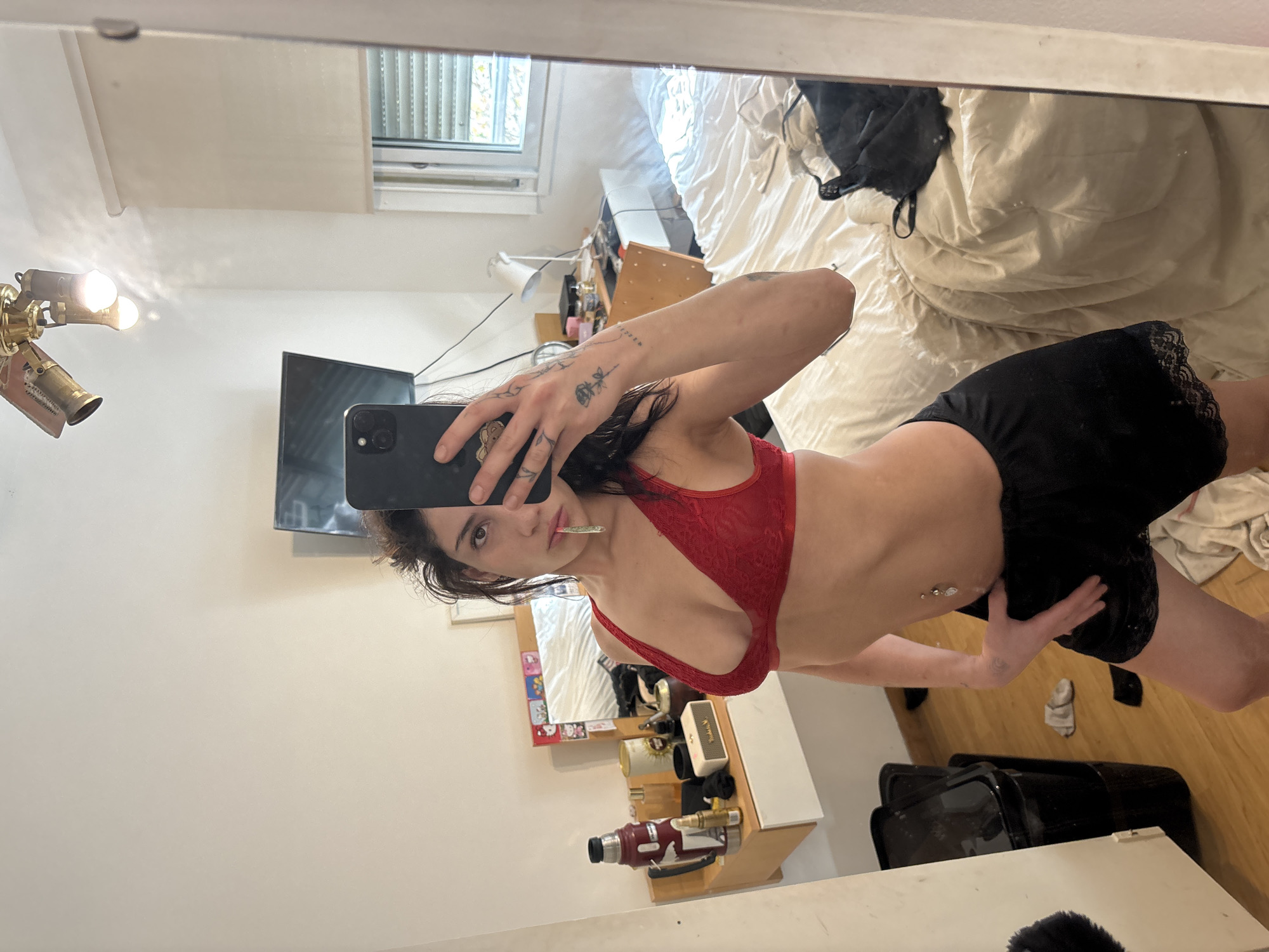 A woman is taking a mirror selfie in what appears to be an apartment. She is wearing a red bra and black shorts. She holds a cigarette in her mouth. There is a bed, a TV, a fan, and a window visible in the reflection. Based on the architectural style and general context, the location is likely Buenos Aires, Argentina.