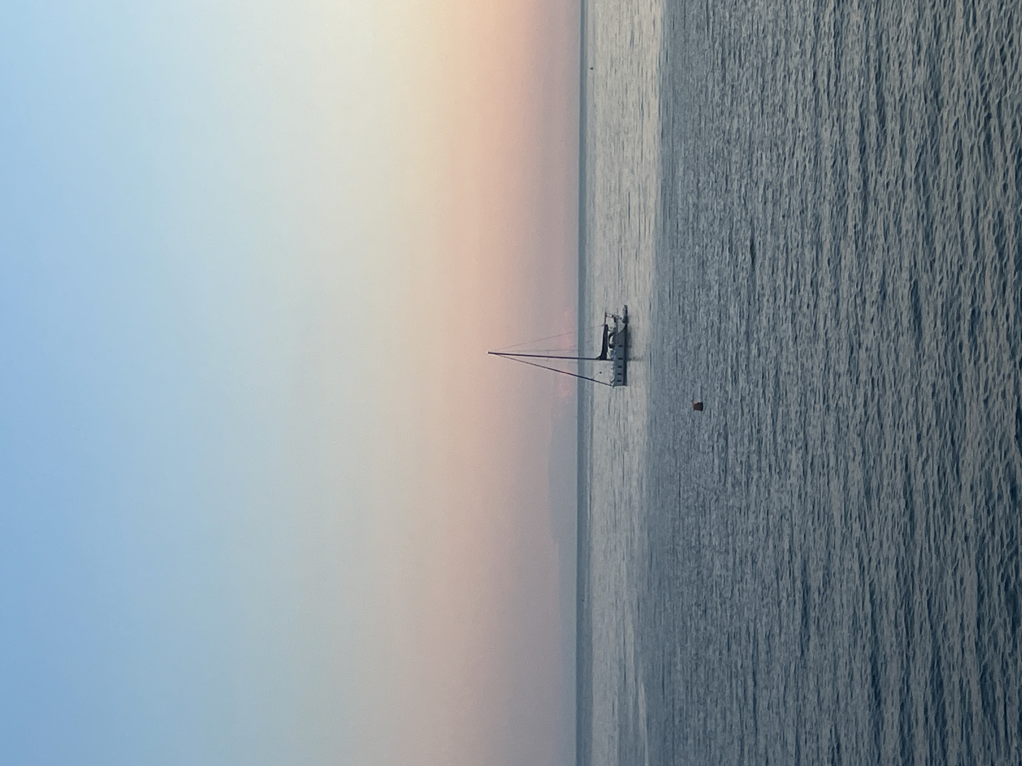 A serene maritime scene captures a sailboat anchored near the city of Budva, Montenegro. The horizon line is visible across the middle of the image with the sea beneath and sky above.
The sailboat is a medium-sized vessel with a white hull and a tall mast reaching upwards. The sails are furled or absent, suggesting it's at rest. Nearby, a small orange buoy bobs gently in the water, likely marking an anchoring spot.
The sea itself is calm, with gentle wave patterns creating a textured surface. The water appears a deep bluish-grey, reflecting the ambient light.
The sky exhibits a gradient of colors, ranging from a light blue at the top to a soft, muted peach and pink towards the horizon. This suggests it could be either dawn or dusk. The light is soft and diffused, lending a peaceful atmosphere to the scene.
The overall composition is simple yet evocative, focusing on the stillness of the water and the quiet solitude of the sailboat under the open sky. The absence of any other vessels or visible structures adds to the sense of tranquility and isolation.