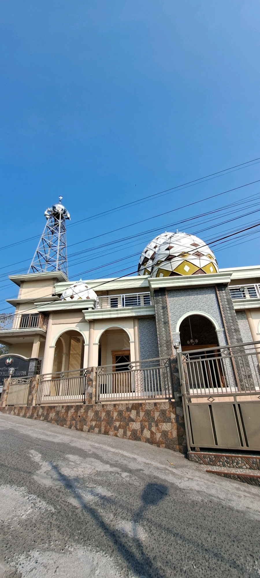 A low-angle, full shot captures a mosque located in Gambiran, Indonesia, under a clear blue sky. The mosque's exterior features a prominent, geometrically patterned dome with white and yellow segments. To the left of the dome, a tall, metallic tower with speakers is visible. The mosque's facade is decorated with arched entrances and adorned with a patterned stone wall at its base. A metal fence encloses the property, and a concrete road leads up to the mosque. Electrical wires traverse the sky above the building. No people are present in the image.