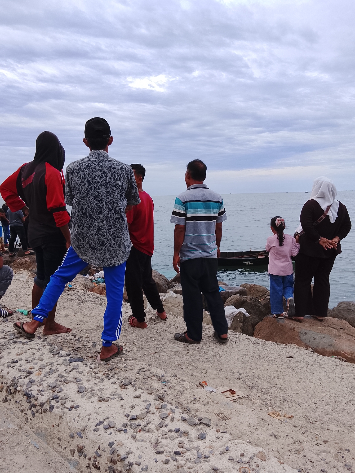 A group of people is standing on a concrete and rock embankment looking out at the ocean under a cloudy sky. The location is Lhokseumawe, Indonesia. From left to right: a person in a black and red hoodie, a person in a patterned shirt and blue pants, a person in a red shirt, a person in a striped shirt and black pants, and a woman in a hijab with a child in a pink shirt. A small boat is visible in the water. There is trash on the embankment.