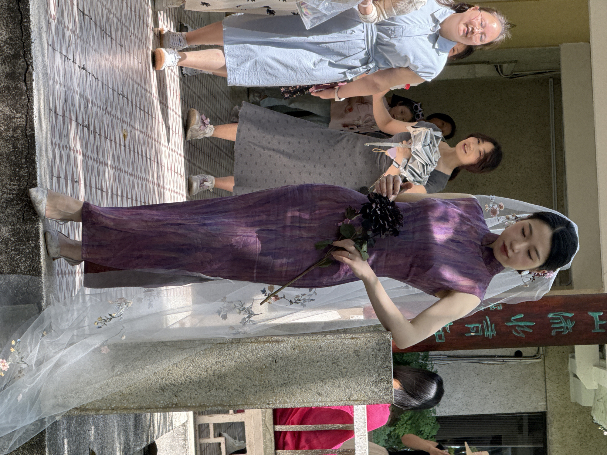 The image depicts a group of people outdoors, possibly in Yuzhong District, China. The primary focus is a woman wearing a purple qipao dress, silver shoes, and a veil. She holds a black flower. Around her, other individuals are present, including a woman in a grey dress, a woman in a blue dress and glasses, and a child. They are standing on a tiled walkway. A sign with Chinese characters is visible in the background. The setting appears to be an urban environment.