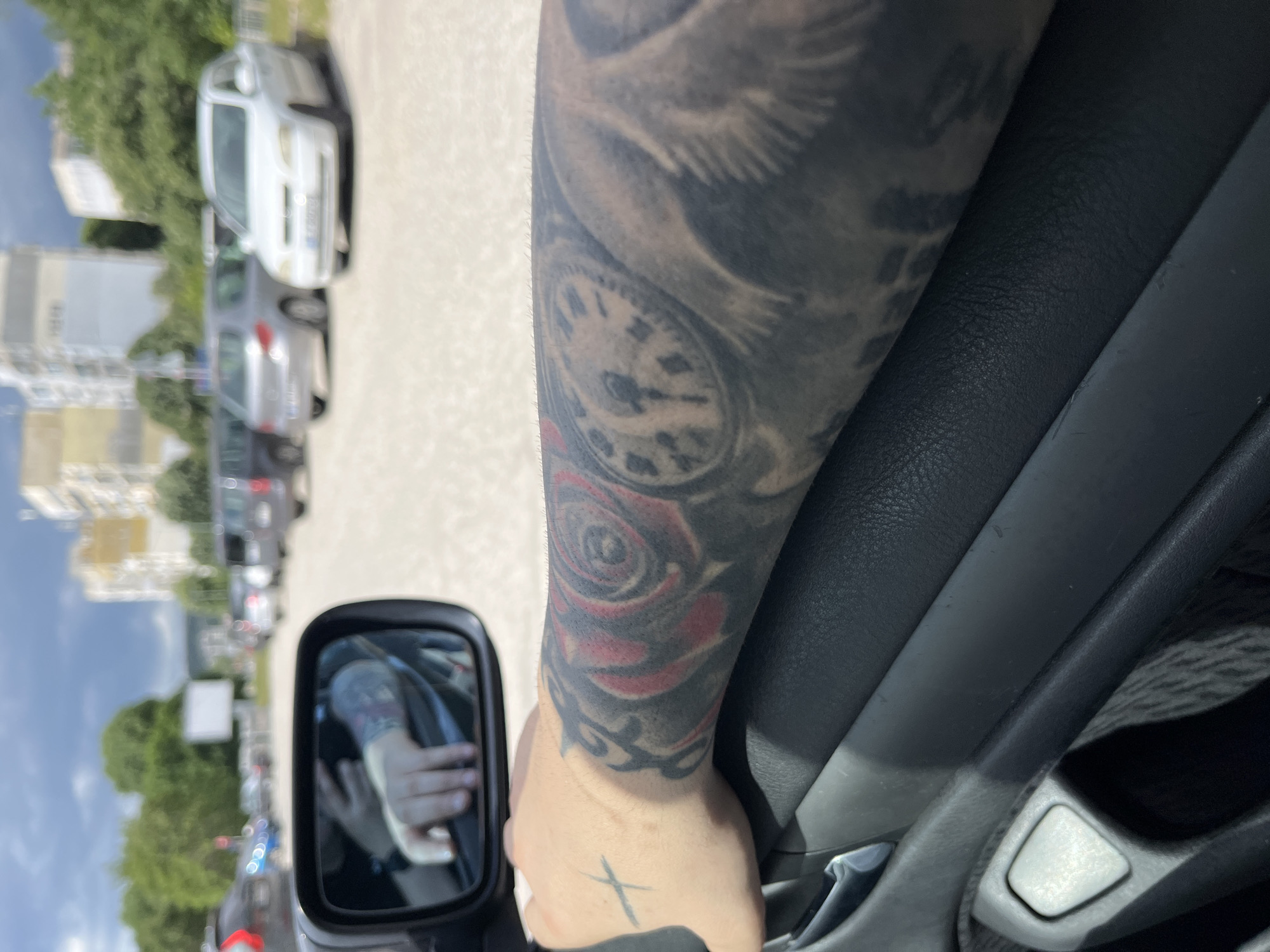 A heavily tattooed arm is shown inside a car, next to the door. The tattoo includes an eagle, a rose, and a clock. The car is parked on a city street in Varna, Bulgaria. Other cars are visible, parked along the street with trees lining the street as well. A side mirror can be seen with a blurred view of the street outside the car.