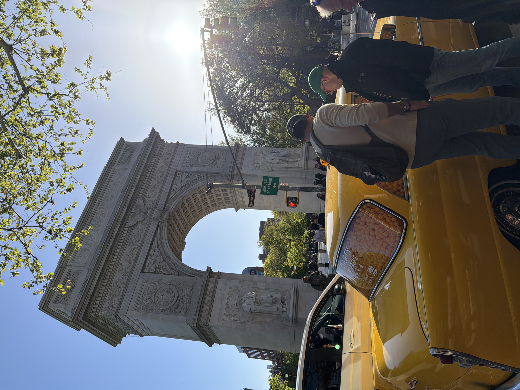 The image features the Washington Square Arch in New York City. The arch is constructed of light-colored stone with intricate carvings and sculptures. In the foreground are people standing near yellow vintage cars. The weather appears sunny and bright, and the sky is a clear blue. The location is clearly in New York City, with the arch serving as a recognizable landmark.