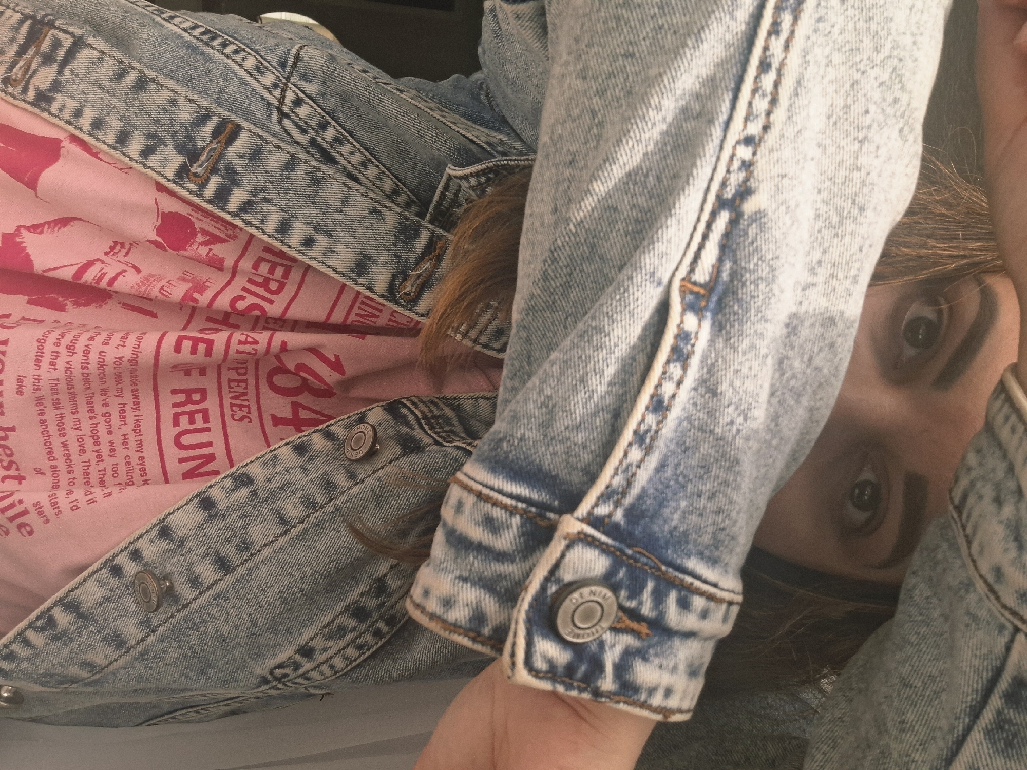 A close-up, slightly tilted shot features a person partially obscured by a light-wash denim jacket. The person's eyes are visible, gazing directly at the camera. They have fair skin and brown hair. A pink t-shirt with text in red is partially visible under the jacket. The text includes "184" and "A1 OPENES REUN." A fragment of the text on the t-shirt includes the phrase "morningsstole away, I kept my eyes." The jacket sleeve's button is branded with the word "DENIM." The location and city of Chaikovsky, Russia, are not evident in the image.