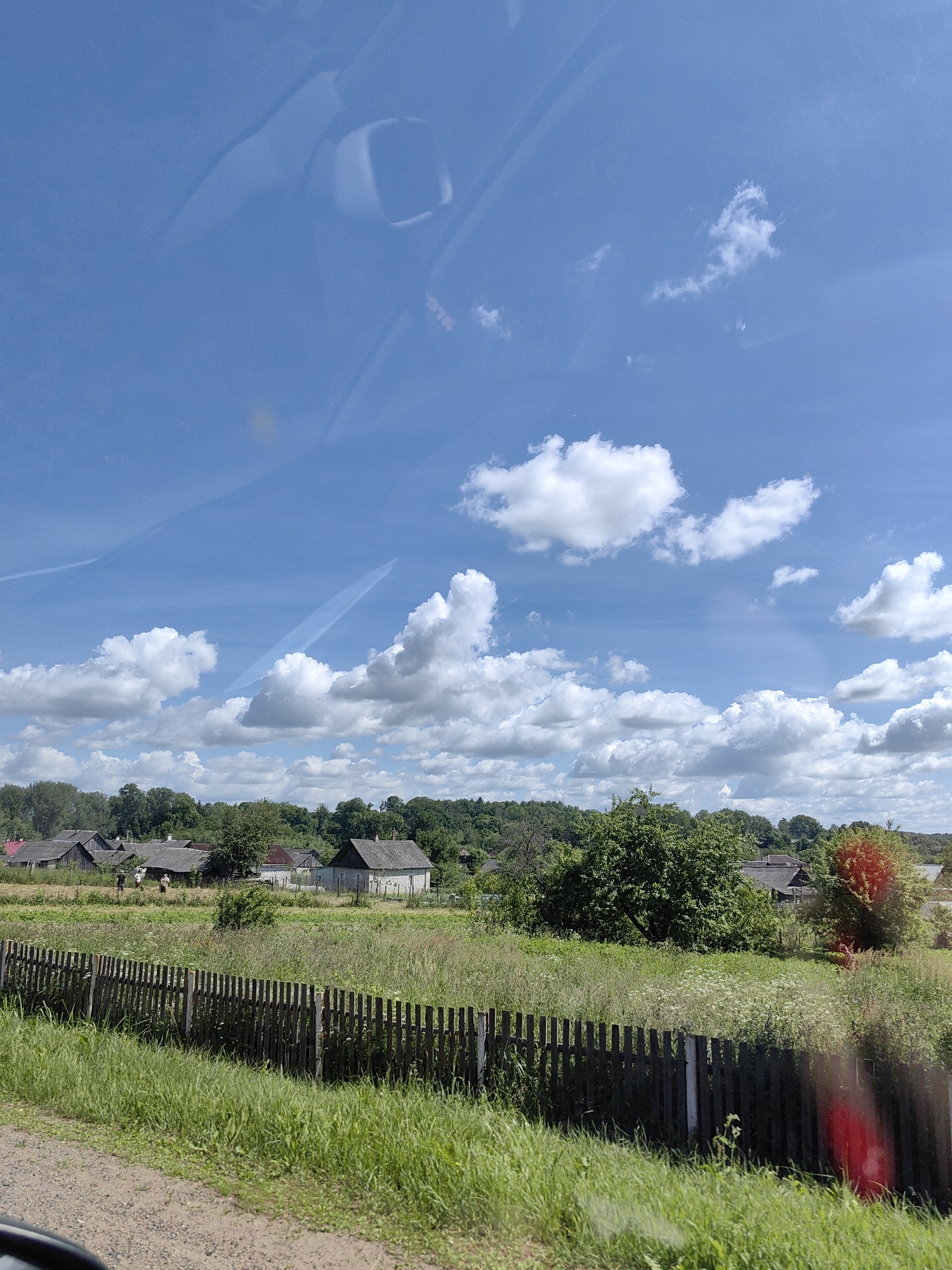 The image depicts a rural village scene under a bright blue sky with scattered white clouds. The foreground features a dirt path bordering a grassy area with a wooden fence running horizontally. Beyond the fence, a field is filled with tall grass and wildflowers, interspersed with several village houses with grey roofs. A dense line of green trees forms the backdrop, suggesting a forested area behind the village. In the distance, more houses are visible, some with chimneys. The sky is a dominant element, with noticeable contrails from aircraft. The scene appears peaceful and idyllic, characteristic of a rural landscape. The location is identified as Ilyanskiy selskiy Sovet, Belarus.
