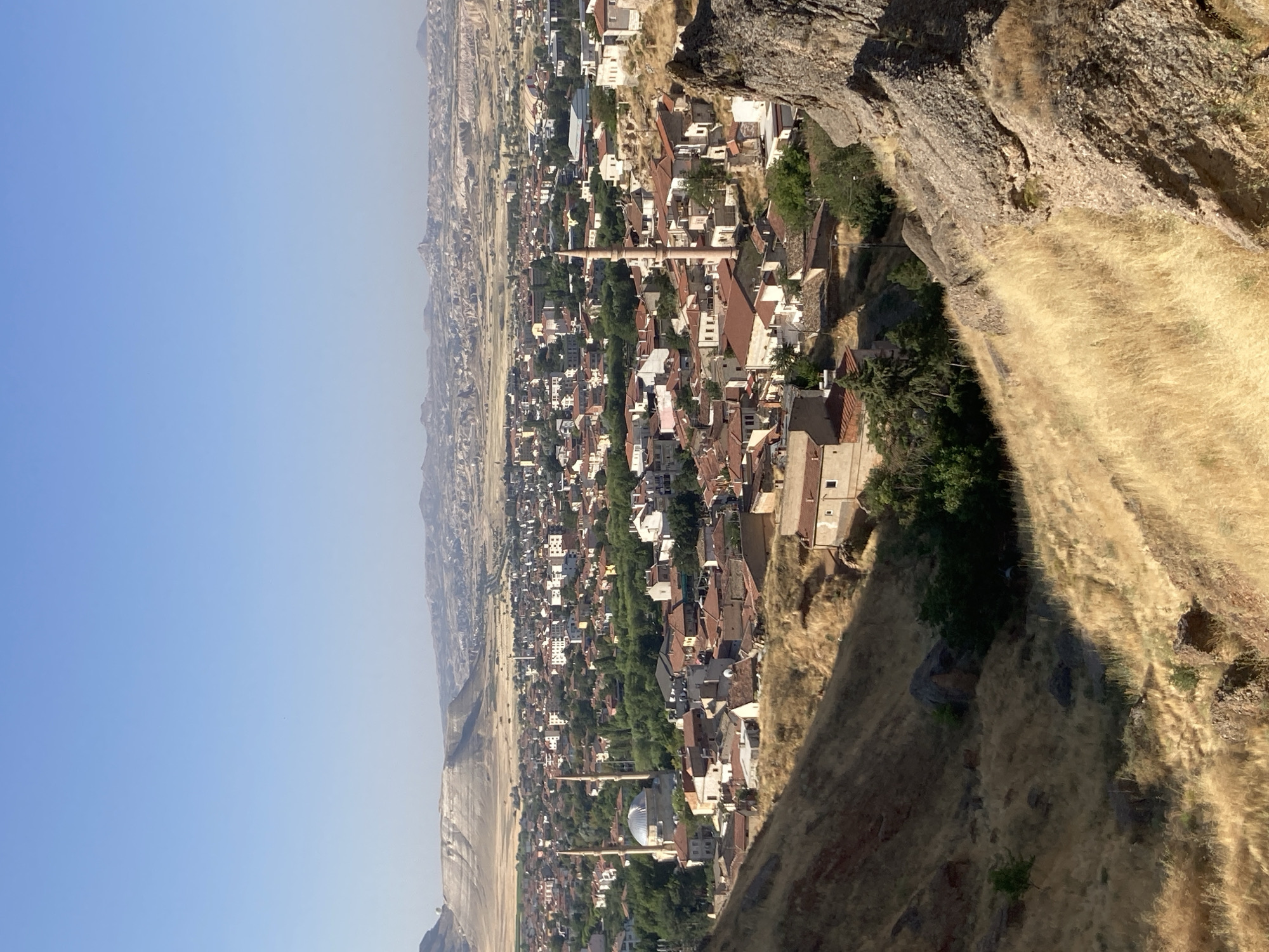The image is an aerial view of Avanos İlçe Merkezi, Turkey, taken on a clear, sunny day. The foreground is dominated by a dry, grassy hillside with rocky outcrops. Below the hill, the city sprawls out, characterized by densely packed buildings with red-tiled roofs. Two prominent minarets, characteristic of mosques, rise above the rooftops. A large mosque with a dome is visible in the mid-ground. The city is surrounded by a dry, arid landscape with rolling hills and distant rock formations, indicative of a semi-arid climate. The sky is a clear, pale blue. There are no people or animals visible in the frame. The overall scene depicts a traditional Anatolian town nestled within a unique geological landscape.
