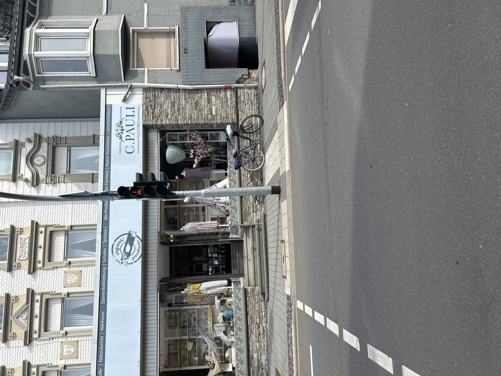 This is an outdoor, eye-level, full-shot photograph taken on a bright, sunny day in the city of Waldbröl, Germany. The scene depicts a street-level view of the facade of several buildings lining a sidewalk. In the foreground, a gray asphalt road with white dashed lane markings is visible, suggesting a typical urban street.
The main focus of the image is a storefront with a large display window, showcasing a variety of items including clothing, home decor, and decorative objects. The store's signage, "C. PAULI NATURE," is prominently displayed above the window. To the left of the storefront, a bicycle with a basket is parked on the sidewalk. A traffic light with a red signal is positioned to the right of the storefront, indicating a stop.
The buildings themselves feature a mix of architectural styles. The building directly behind the storefront appears to be older, with decorative window frames and a white brick facade. Adjacent to it, on the right, is a more modern building with a gray stucco exterior and bay windows. A building number, "55," is visible on the gray facade. The sidewalk is paved with gray tiles and stone. The environment is clean and orderly, with no visible people in the frame. The overall impression is one of a quiet, well-maintained commercial street in a European town.