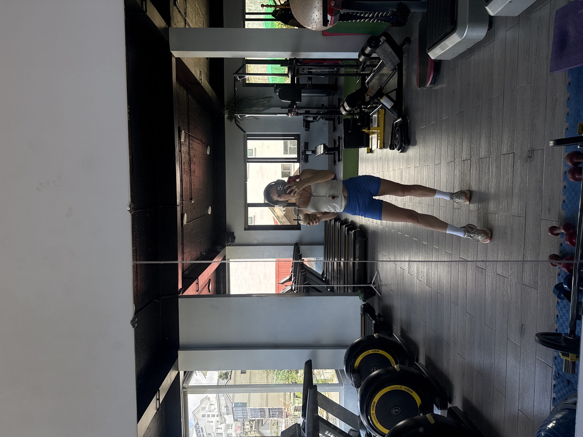 A person is reflected in a mirror in a gym. They are standing and holding a drink. The person is wearing headphones, a white tank top, blue shorts, white socks, and athletic shoes. The gym is equipped with treadmills, weight machines, and free weights. The gym has large windows that look out onto a city scene, with buildings and trees visible. The floor is tiled. The ceiling is black.