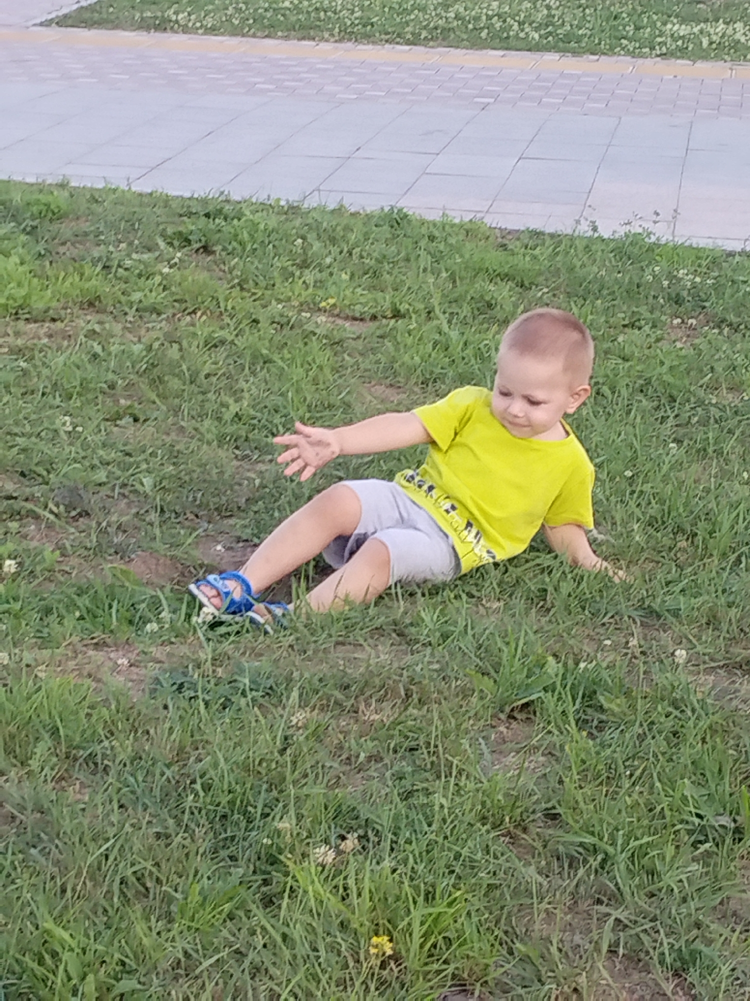 A young boy, appearing to be around two years old, sits on the grass in a park, his body angled towards the right of the frame. He is wearing a short-sleeved neon yellow t-shirt and light gray shorts. His right arm is extended outward and slightly upward, with his hand open and fingers spread, as if reaching for something. He wears blue sandals with white accents on his feet. The boy's head is turned to his left, and he appears to be looking down with a focused expression. The ground beneath him is covered in green grass, with patches of bare earth visible. In the background, a paved walkway made of gray square tiles with a yellow border runs horizontally across the upper portion of the image, suggesting an urban park setting within Komsomolsk-on-Amur, Russia. The overall scene captures a moment of a child exploring his surroundings in an outdoor environment.
