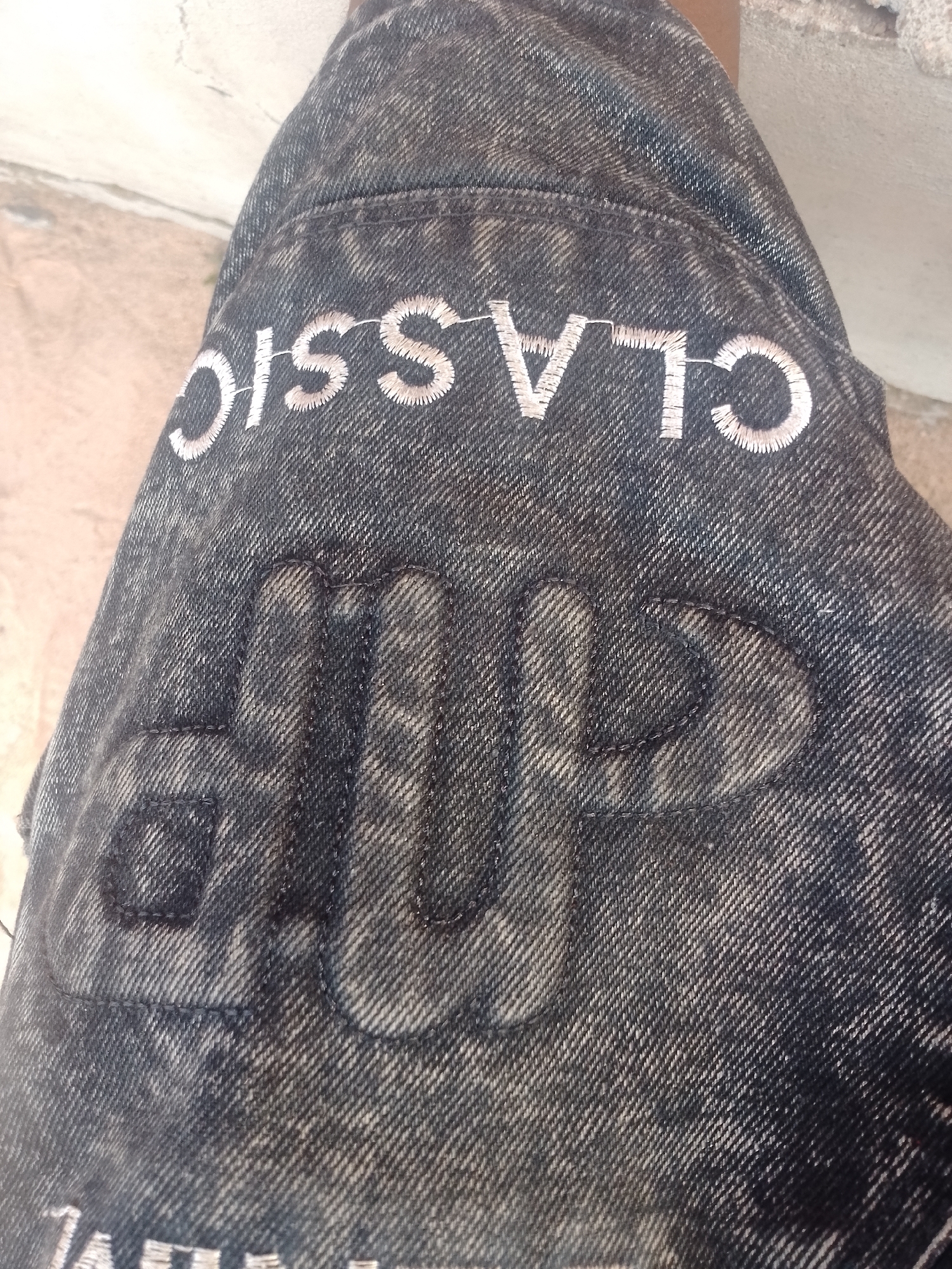 A close-up, angled shot shows a dark-washed denim fabric with embroidered lettering and graphics. The lettering "CLASSIC" is embroidered in white thread across the upper portion of the frame, with each letter outlined in a thin, lighter thread. Below the "CLASSIC" text, there is a large, raised graphic composed of interconnected, puffy letters that appear to spell "DUP". This graphic is also embroidered with dark thread, giving it a slightly textured and shadowed appearance against the denim. The denim itself has a slightly faded, acid-washed look, with lighter streaks and darker areas. The background is out of focus but appears to be a textured, light-colored surface, possibly a wall or ground.
