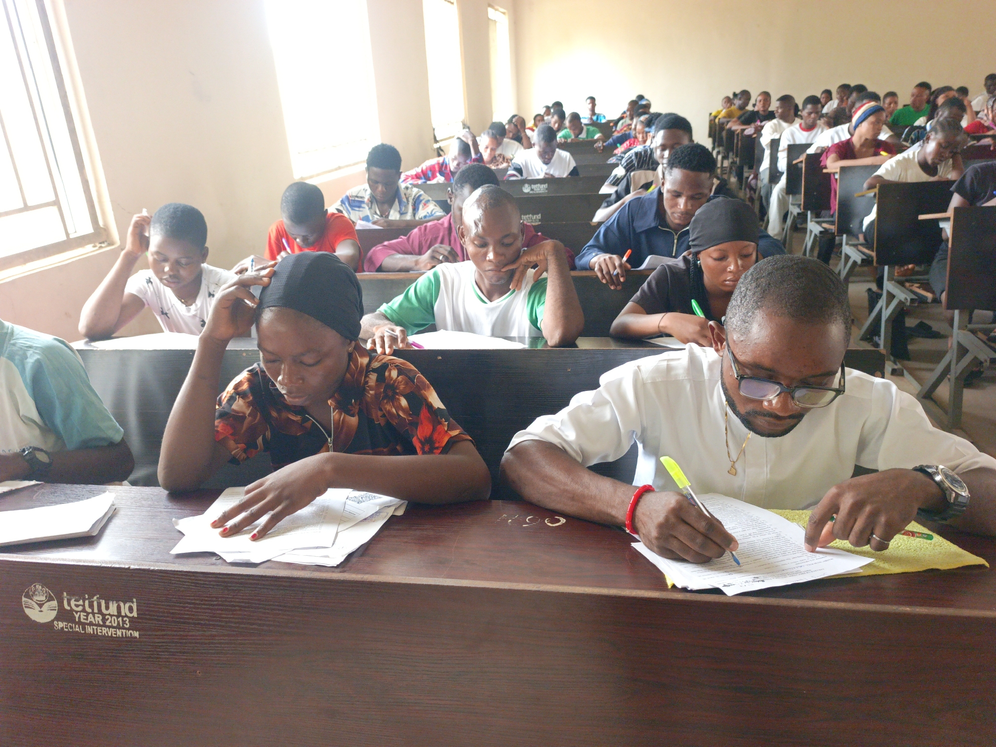 **Location:** Classroom in Akwanga, Nigeria.
**Scene:** A classroom filled with students taking a test or exam.
**People:** The image shows a large group of students, likely university or higher education level, seated at desks in rows. They are of various ages and are likely all Nigerians. They appear to be concentrated on writing or reading papers. One man in the foreground appears to be a proctor or possibly a professor/teacher observing the students.
**Subjects:** Students are engaged in taking a test/exam.
**Objects:** There are papers on desks (presumably exam papers), pens/pencils, desks, chairs arranged in rows. The desks in the front row has a Teitfund Year 2013 Special Intervention logo on it. One person has a wristwatch.
**Actions:** Students are writing, reading, concentrating. The instructor is writing, grading, or making notes on paper.
**Interactions:** Primary interaction between each student and the written material.
**Environment:** The classroom is a fairly standard room. There are rows of desks and chairs. Light is coming through windows, indicating it is daytime. The classroom's general appearance suggests a basic educational facility.