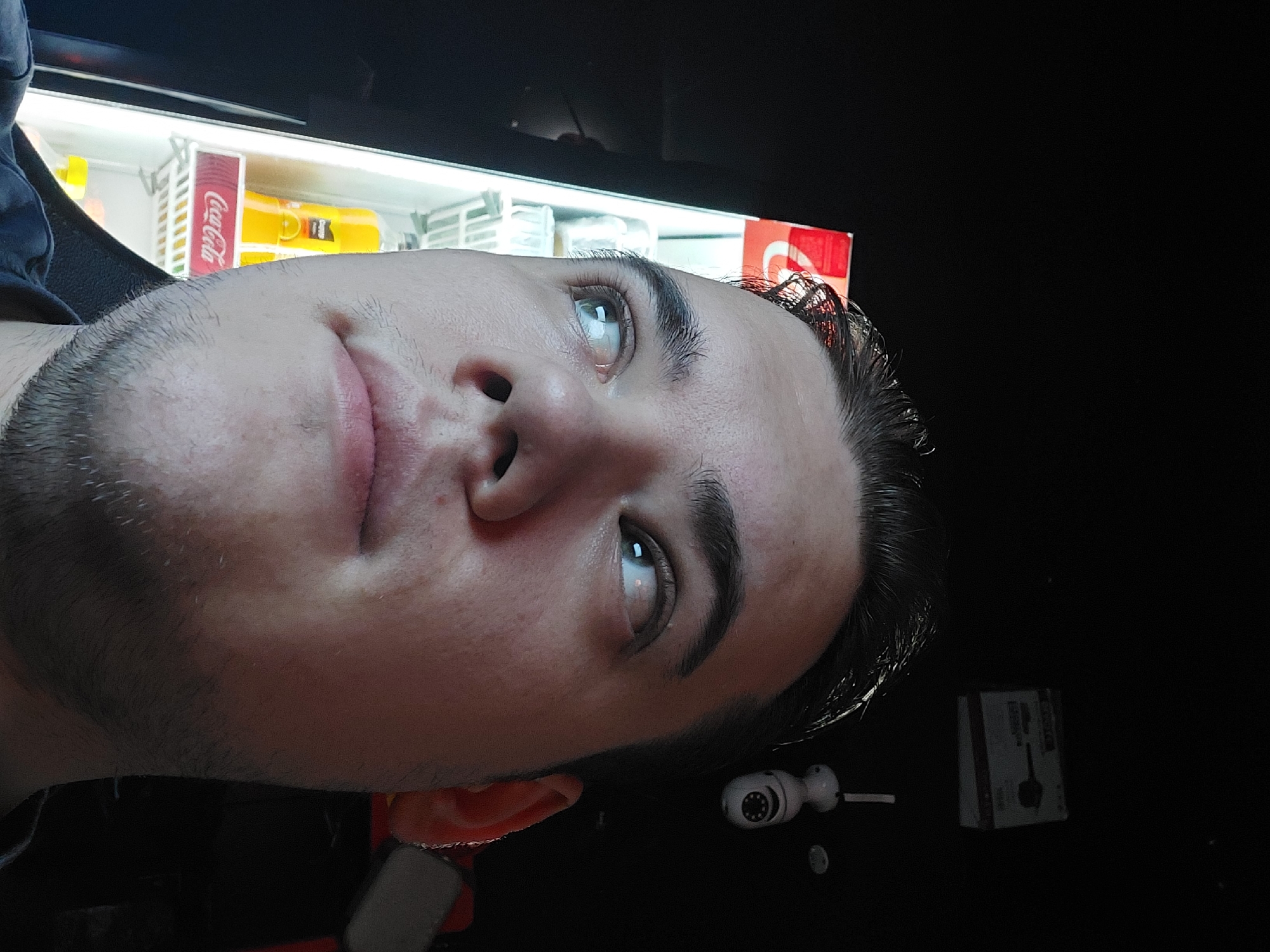 A close-up, tilted image focuses on a man's face, with his head angled towards the left side of the frame. He has light skin, dark hair, stubble, and his eyes are looking upwards. Behind him, a cooler with visible Coca-Cola branding and other beverages is discernible. In the lower right corner, objects including a small white surveillance camera and a box-shaped item are visible against a dark background. The image was taken in the city of Tashkent, Uzbekistan.