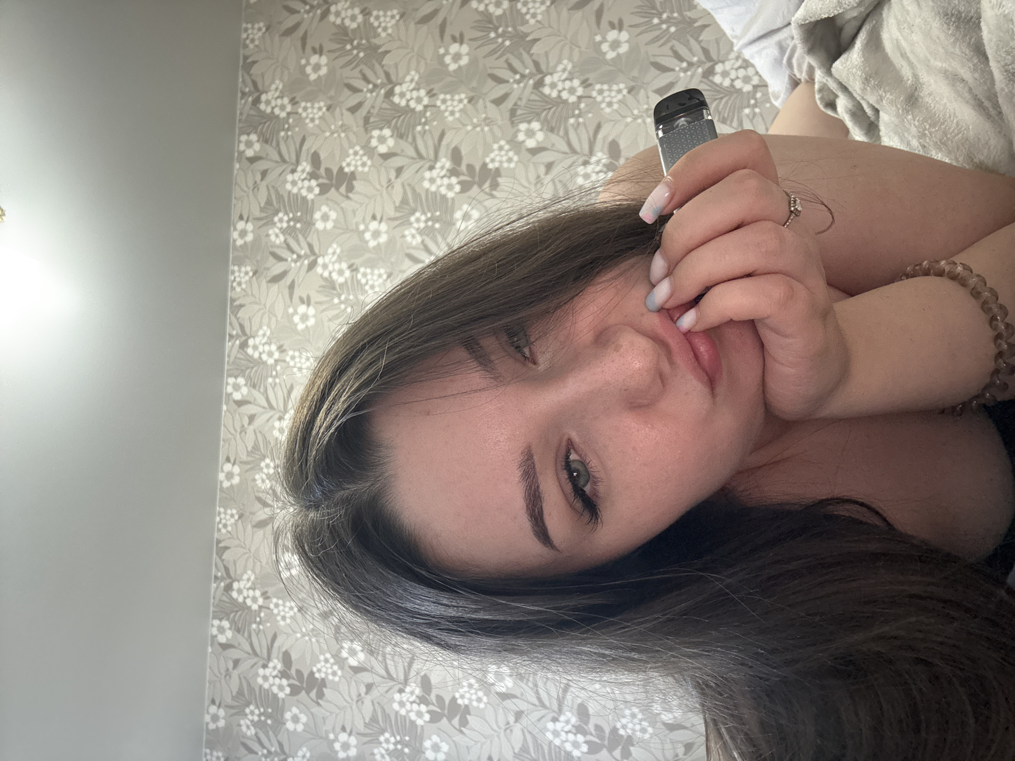 The image is a close-up, rotated portrait of a young woman lying down. She is facing the camera, with her head tilted. Her eyes are open and looking directly at the viewer. Her hair is dark brown and falls around her face. She is wearing a ring on her right hand, which is near her face. She is also holding a small electronic device, possibly a vape, in her right hand. Her nails are painted in a gradient of white, pink, and blue. A beaded bracelet is visible on her left wrist. She appears to be in a bedroom, with floral wallpaper in the background and a light-colored blanket or duvet visible in the upper right corner. The lighting suggests it is daytime, likely indoors. The overall mood conveyed is relaxed and intimate. There is no visible text in the image.