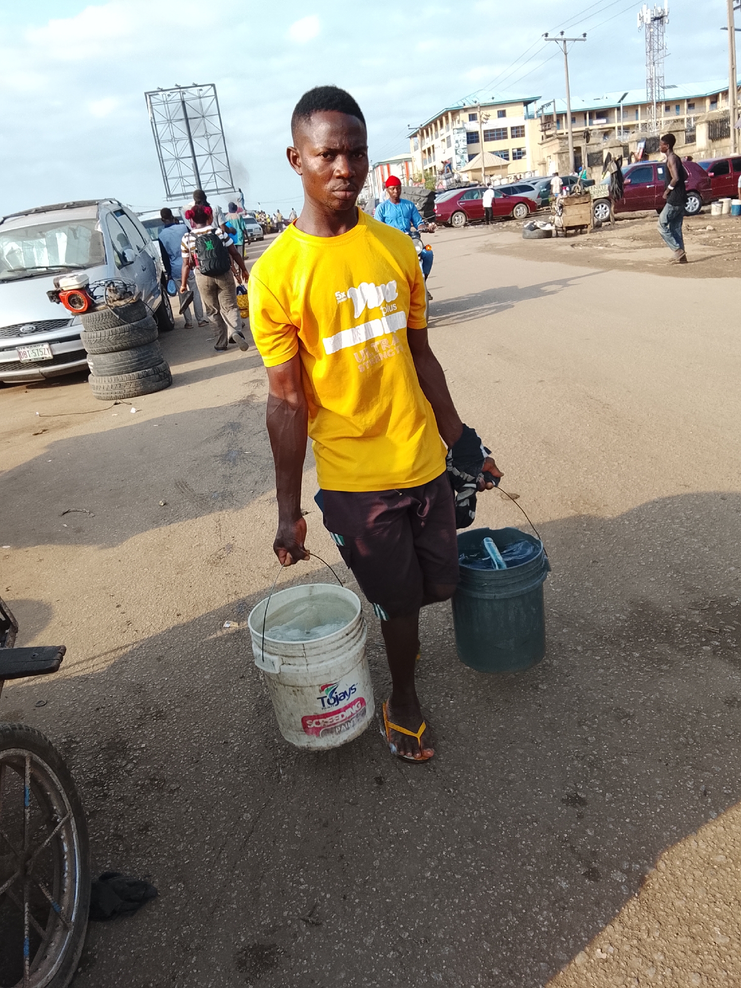 A young man stands in the middle of a street, holding a bucket in each hand. He wears a yellow t-shirt, brown shorts, and yellow sandals. The buckets appear to contain water.
In the background, there are several cars, pedestrians, and buildings. A billboard structure is visible in the distance. A stack of tires sits next to a silver car.
The scene is likely located in a busy urban area in Abuja, Nigeria.