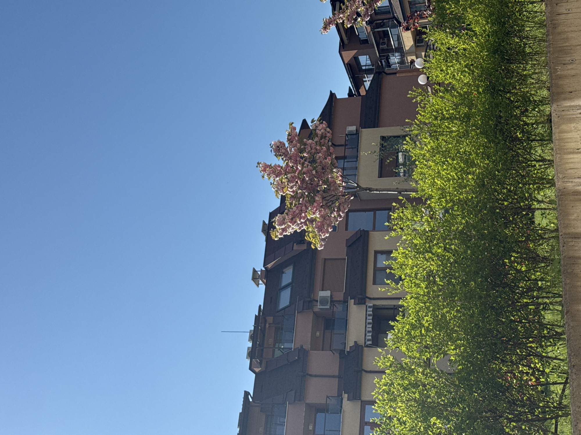 A low-angle, daytime shot depicts an apartment building and trees against a clear blue sky. The building is multi-story, with an architectural style featuring angular projections and a mix of brown and beige exterior finishes. It includes windows, balconies, and what appear to be air conditioning units attached to the exterior. A blossoming tree with pink flowers is partially visible in front of the building. Green trees with leaves are situated along the right side. The image may be of Sofia, Bulgaria.