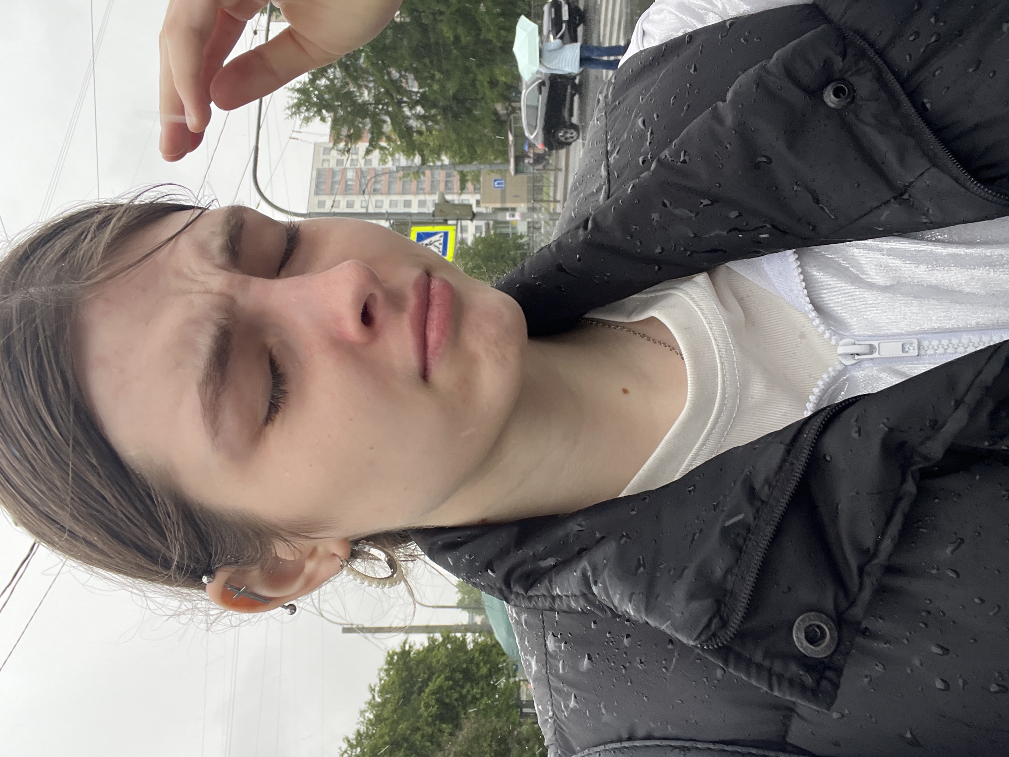 A medium, close-up shot depicts a person with their eyes closed, wearing a white shirt and a black puffer jacket, visibly wet with raindrops. The person's face is angled upwards slightly. They have several piercings on their left ear. A hand is positioned above their forehead. In the background, out of focus, is an urban scene. There are buildings, trees, and road signs visible. A pedestrian crossing sign with a yellow and blue triangular emblem is observable. There are cars and people crossing the street, one person with an umbrella. The environment is consistent with a rainy day in a city. The location is Saint Petersburg, Russia.