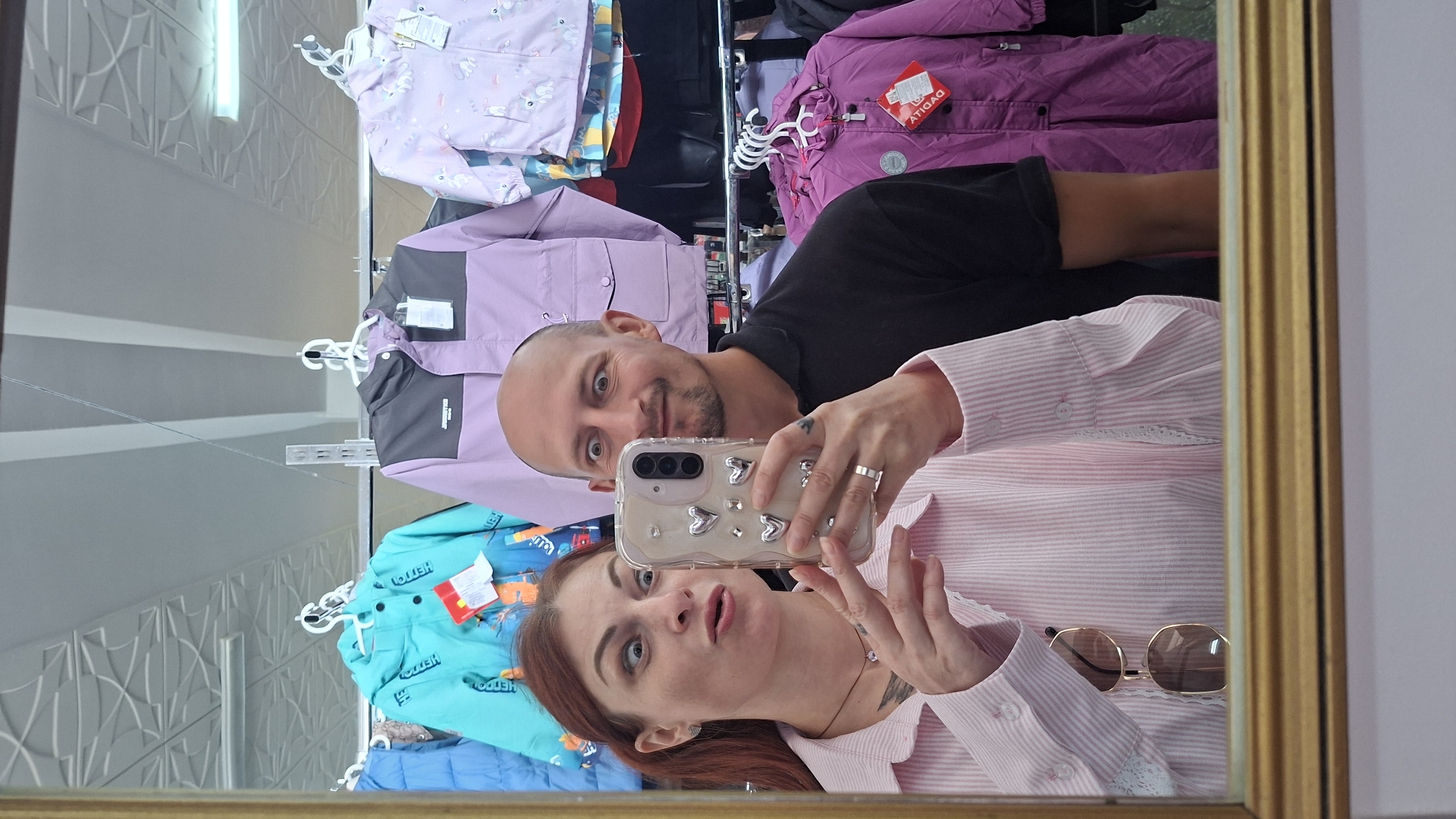 A man and a woman are taking a selfie in a store. The woman is on the left, wearing a pink and white striped shirt and sunglasses hanging from her shirt. She has reddish-brown hair pulled back and is holding a smartphone with a heart-shaped decoration in her right hand. Her eyes are wide and her mouth is open in a surprised expression. The man is on the right, wearing a black polo shirt. He is bald and has a goatee. His eyes are also wide and he is smiling. Behind them, children's jackets are hanging on display racks. The store appears to be a clothing store. The image was taken in Myadzyel, Belarus.