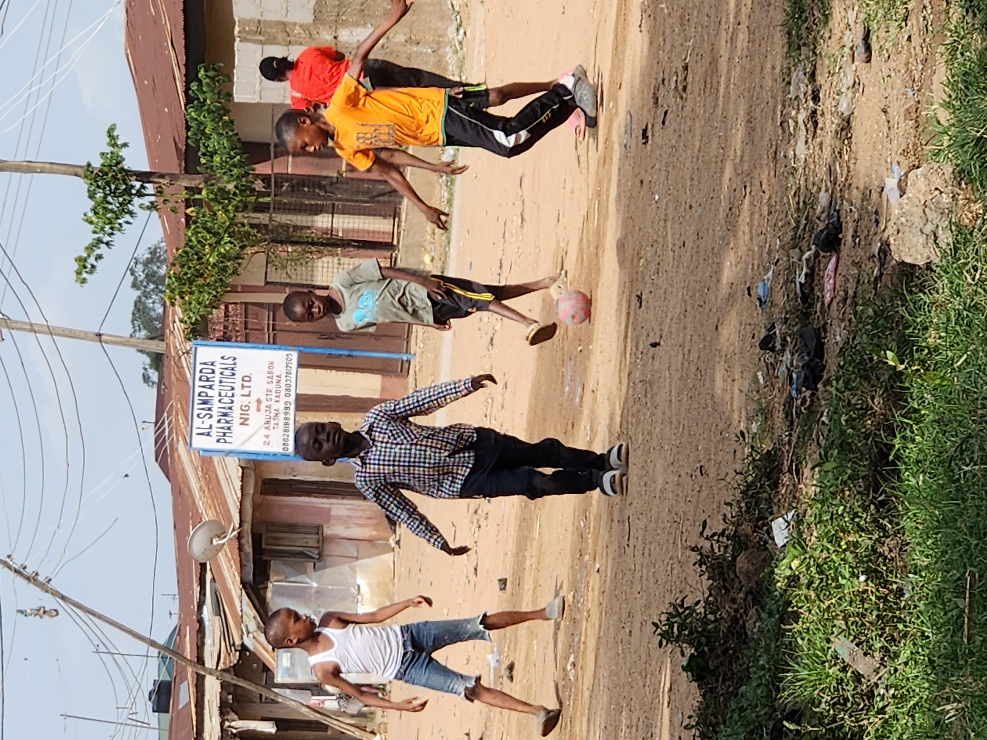The media file shows a group of young boys playing with a soccer ball in a dusty street, likely in Sabon Tasha, Nigeria. The scene is set outdoors, with a dry, dirt road surface and some sparse green vegetation on the right side. In the background, there are structures that appear to be buildings with corrugated metal roofs, suggesting a somewhat informal or rural setting. The sky is visible and appears bright, indicating daylight, possibly late morning or afternoon.
Several boys are actively engaged in the game. One boy in an orange shirt is kicking the ball, his body contorted in mid-action. Another boy in a green t-shirt is near the ball, and a third boy in a plaid shirt is also in motion, possibly reaching for the ball. Another boy wearing a white tank top and denim shorts is also present, observing or participating in the game. Their postures and expressions convey a sense of energetic play and youthful engagement.
Visible text on a signpost provides context, identifying "AL-SAMPARDA PHARMACEUTICALS NIG. LTD." and an address in "SABON TASHA KADUNA," along with phone numbers. This confirms the location as Sabon Tasha, Kaduna, Nigeria. The overall impression is one of a lively street scene with children enjoying a game of soccer in a typical urban or semi-urban environment in Nigeria. The lighting suggests a sunny day.