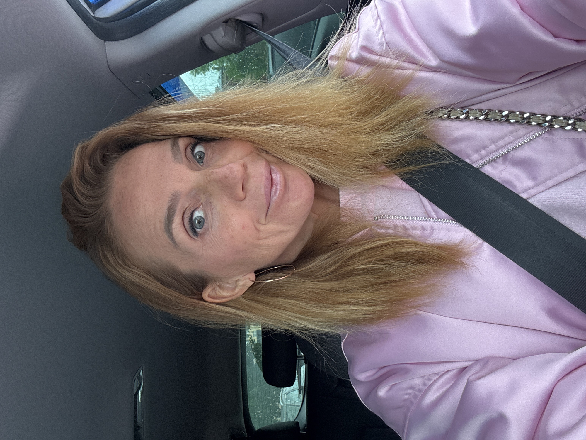 A woman with blonde hair, wearing a pink jacket and a seatbelt, is in a car. She is wearing a gold hoop earring in her left ear. The environment appears to be the interior of a vehicle, with the city visible through the window. The location in the city of Saint Petersburg, Russia is not discernible from the image.