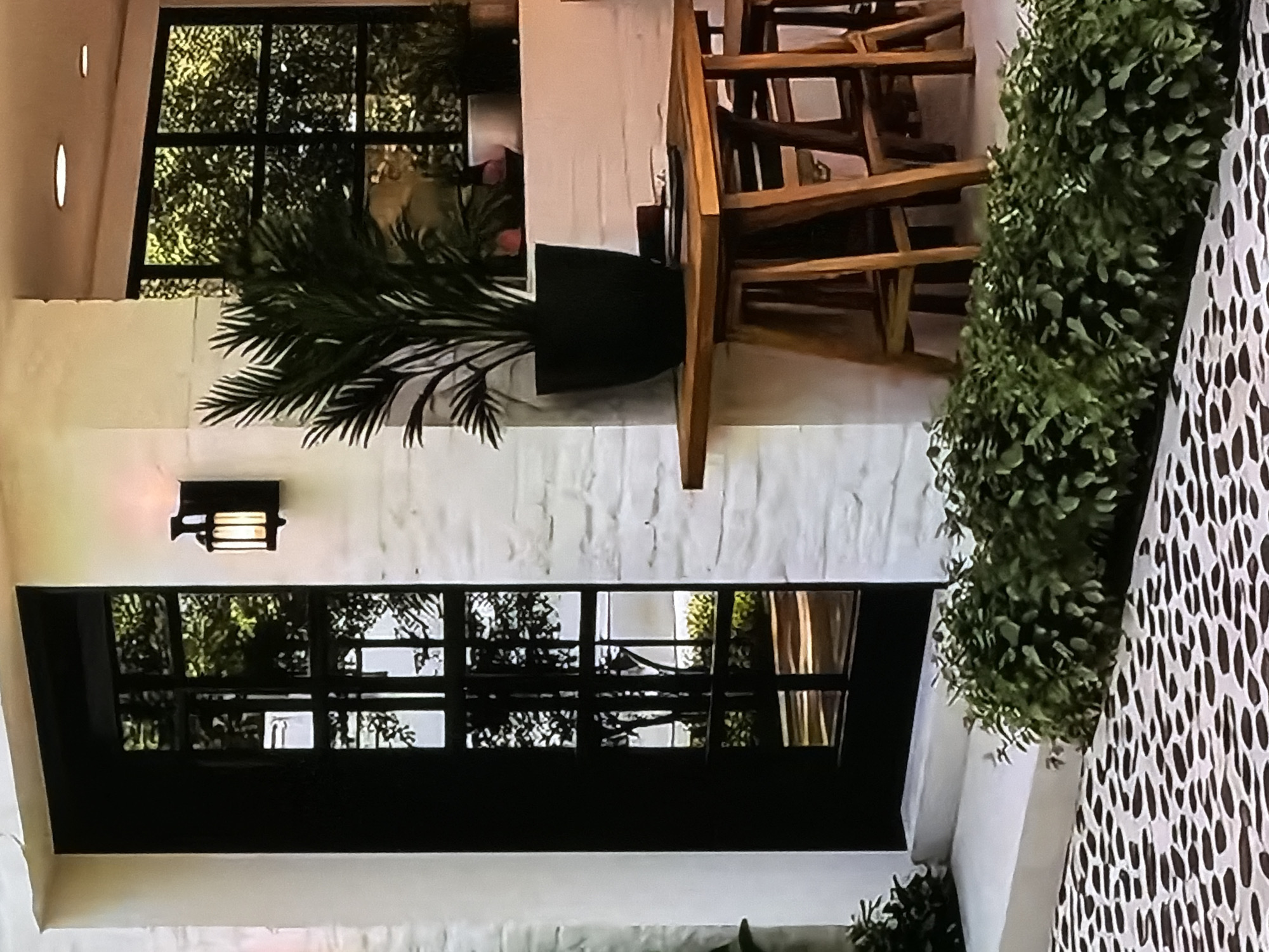 This image captures a chic, possibly semi-outdoor, area in Sofiivska Borshchahivka, Ukraine, perhaps a restaurant patio or an upscale residential space, seen under atmospheric lighting. The scene appears to be set in the evening or night, indicated by the warm glow emanating from a wall-mounted light fixture.
To the left, a prominent black-framed window, divided into multiple panes, reflects lush green foliage from outside. Below this, a section of textured, whitewashed wall is visible. Centrally, a large potted plant with dark green, palm-like leaves is housed in a black planter, positioned on a white ledge or floor. Above a lower, similar black-framed window or door section, a black wall sconce casts a warm, inviting light onto the textured wall, which appears to be whitewashed brick or stucco.
To the right of the potted plant, a light wooden table stands, upon which several matching wooden chairs are neatly stacked. This arrangement suggests the area is either closed, being prepared for opening, or undergoing cleaning. Further to the right, a long, dense arrangement of green foliage, possibly artificial boxwood or similar plants, lines a raised edge or planter box. To the far right, a decorative screen or partition features an abstract, organic pattern of white shapes against a darker background, adding a stylish element to the space. No people are visible, and there is no discernible text within the image.