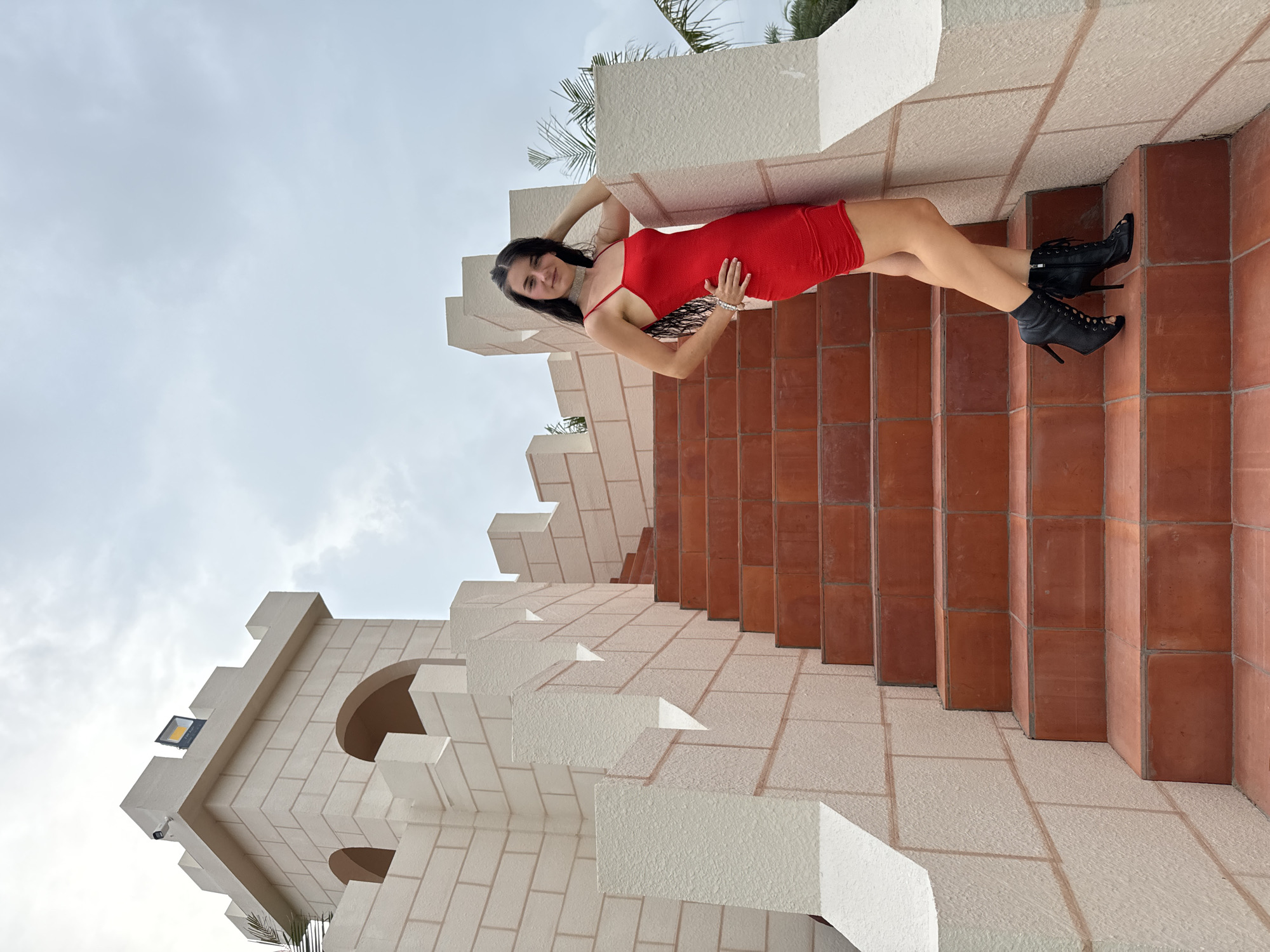 A young woman is captured posing on an outdoor architectural structure in Mui Ne, Vietnam. She has long, dark hair and is dressed in a bright red, form-fitting mini-dress, complemented by black open-toed high-heeled ankle boots, a silver choker necklace, and a pearl bracelet. She is lying on her back across a series of light-colored, textured architectural blocks that resemble castle battlements or stepped terraces. Her head and upper body are slightly elevated, with her right arm casually behind her head and her left hand resting on her stomach. She smiles confidently towards the camera.
The structure features contrasting reddish-brown tiled stairs and wall sections. Hints of green palm fronds are visible in the background, suggesting a tropical or resort environment. The sky is overcast, indicating daytime conditions with diffused light. The architecture itself is distinctive, with angular blocks and rounded archways forming what appears to be a multi-level building. A small light fixture and a security camera are visible on one of the white block sections in the background. The overall scene depicts a fashionable individual striking a pose within an interesting and unique architectural setting. No visible text is present.