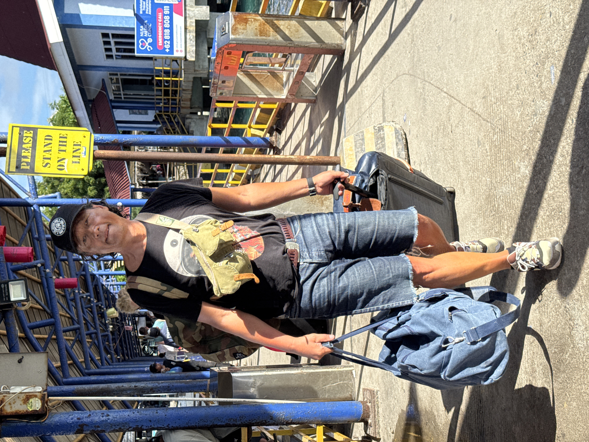 A man stands smiling with his luggage in an outdoor waiting area, likely a pier or ferry terminal, under bright daylight. He is positioned next to a blue metal railing, with strong shadows cast across the concrete ground. The man, who appears to be an adult, wears a black cap, black t-shirt, denim shorts, and sneakers. He has an olive green cross-body bag and a camouflage-patterned item on his back, and holds the handle of a black rolling suitcase with one hand and a blue duffel bag with the other.
In the mid-ground, a prominent yellow sign reads "PLEASE STAND ON THE LINE," suggesting he is queuing or waiting to board. The background shows parts of a building, some industrial-looking metal structures (possibly part of a loading ramp), and other people in the distance, including one lying down. Above, a blue and white billboard is visible for "NUSA MEDICA CLINIC," displaying "Worldwide Travel Insurance Assistance" and an "EMERGENCY CALL +62 818 808 911." The "+62" country code strongly indicates the location is in Indonesia. The scene captures a moment of travel, possibly as the man prepares for a journey by sea.