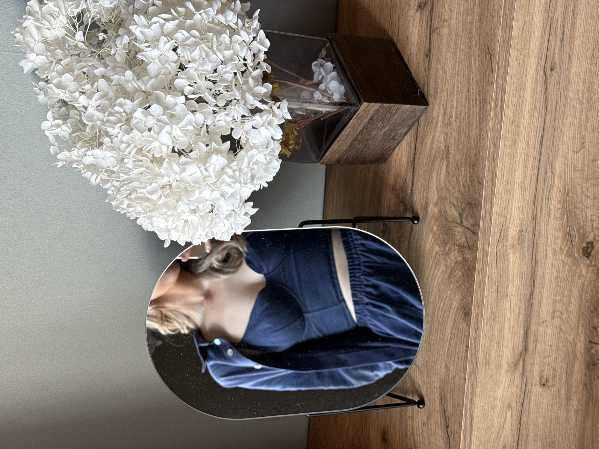 The image shows a partial reflection of a woman in a mirror. She is wearing a dark blue top with a fitted bodice and a button-down portion. The mirror is oval-shaped and has a black stand. To the left of the mirror is a large bouquet of white flowers, possibly hydrangeas. Behind the flowers is a gray wall. To the right of the mirror is a wooden vase and a wood floor. The location is Chaikovsky, Russia.
