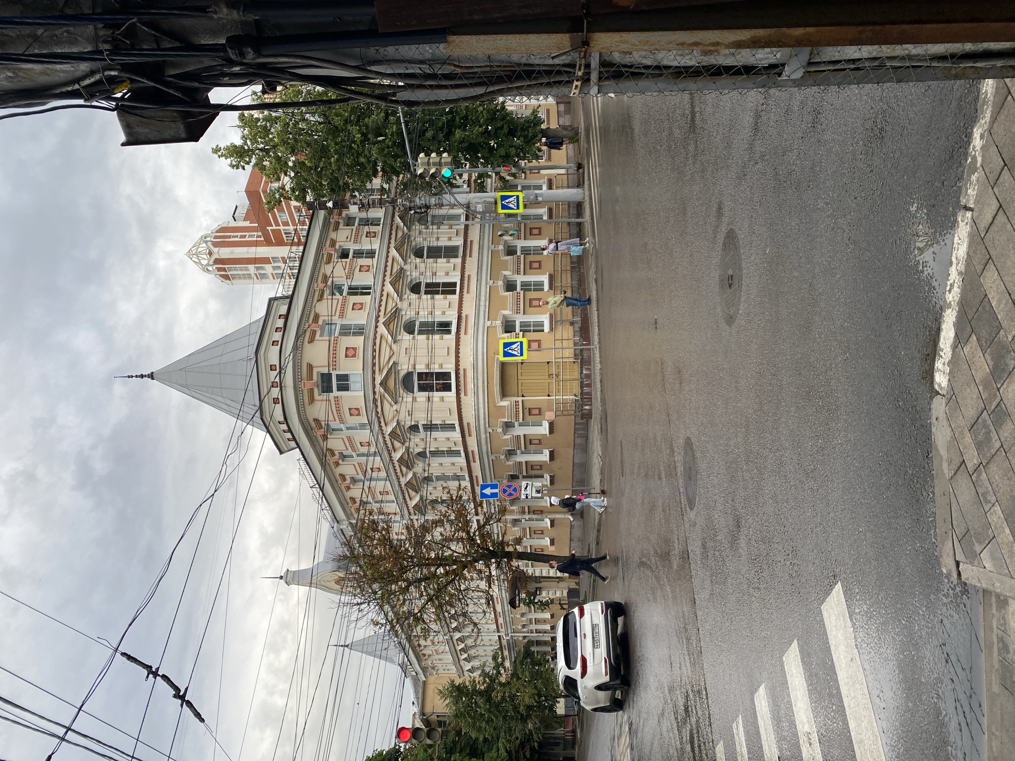 The image captures a street scene in Voronezh, Russia, under an overcast sky, suggesting a cool or rainy day. The wet asphalt of the road and the puddles indicate recent precipitation. Several people are visible, some crossing the street at a marked crosswalk, others walking along the sidewalk. A white SUV is parked on the right side of the road.
The architecture is a prominent feature, with a striking yellow and cream-colored building dominating the left side of the frame. It has a detailed facade with ornate trim, arched windows, and a distinctive pointed spire-like roof structure. Further in the background, a taller, more modern building with red brick and white accents can be seen. Utility wires are strung across the upper part of the image, adding to the urban feel.
Traffic signals are visible, with a green light illuminated, and pedestrian crossing signs are posted. One sign shows a pedestrian figure within a blue diamond, indicating a pedestrian crossing. A blue directional sign with a white arrow points left.
A tree with sparse autumn foliage stands near the building and crosswalk, its branches bare in some areas. The overall mood of the image is somewhat muted, consistent with the cloudy weather and the urban environment. The license plate of the white SUV is partially visible but not legible.
